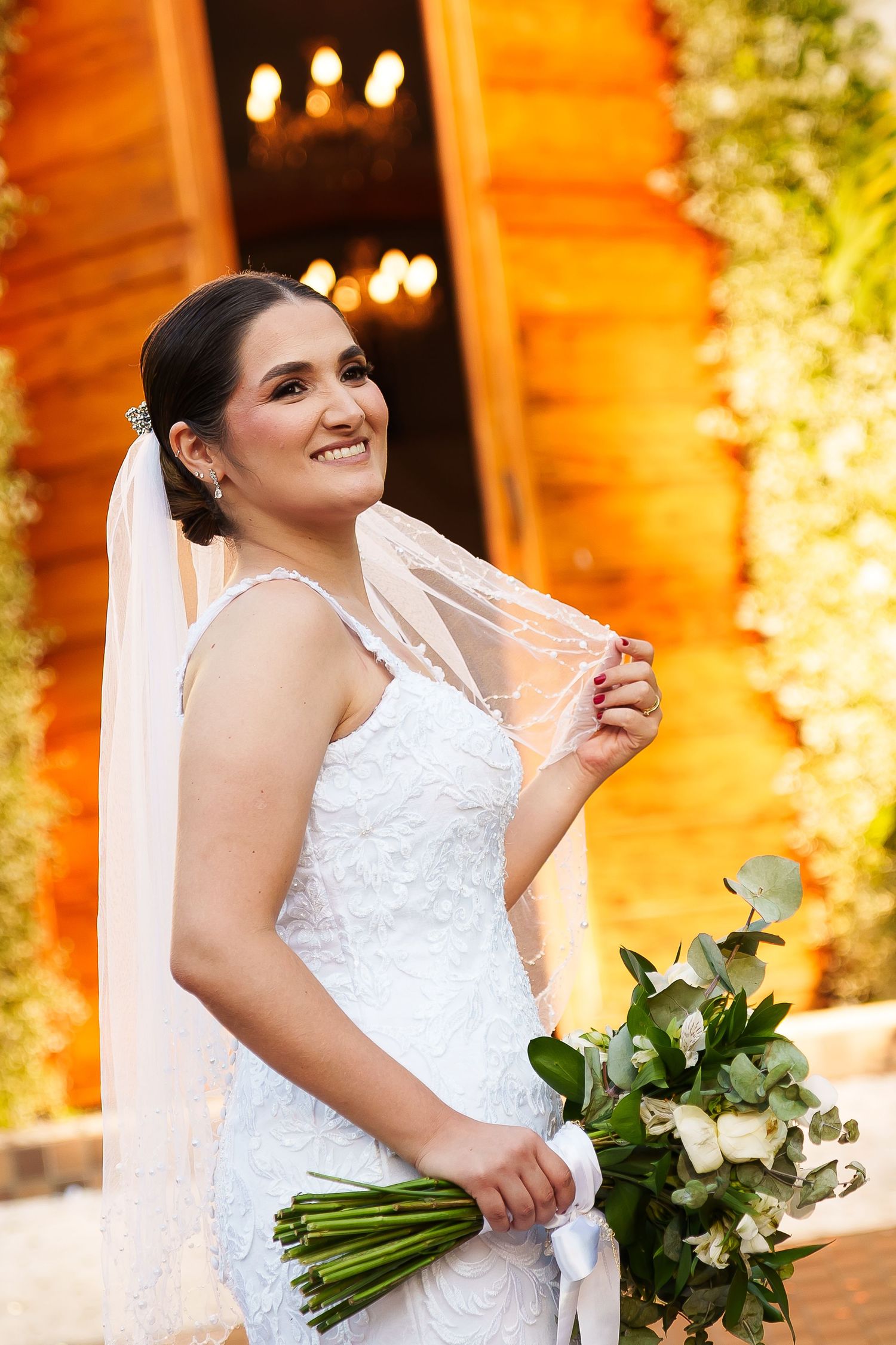 Smiling bride in white lace gown holding bouquet of white roses and greenery, wearing veil outdoors near warmly lit venue.