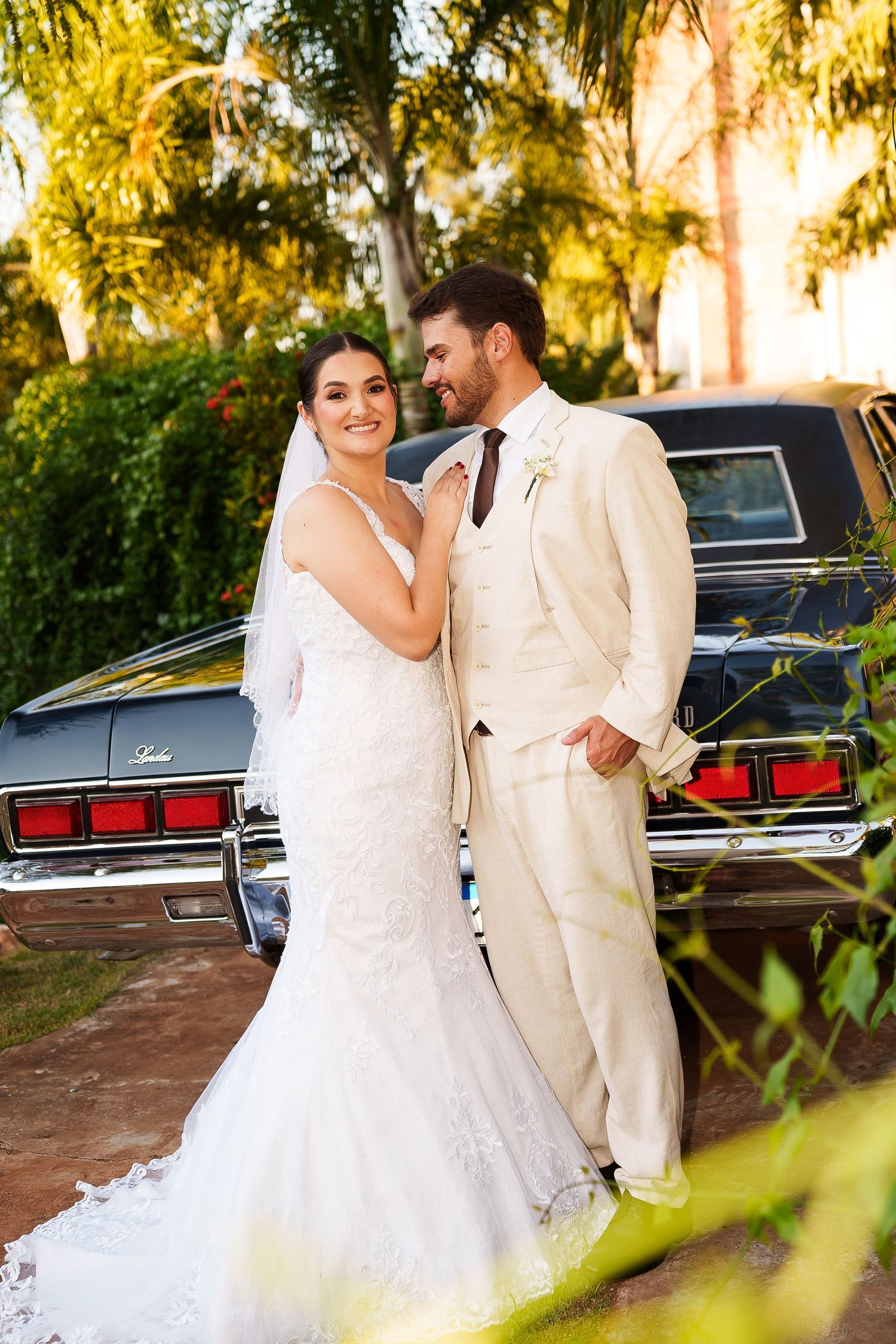 Bride and groom pose together in front of a vintage black car surrounded by lush greenery on their wedding day.