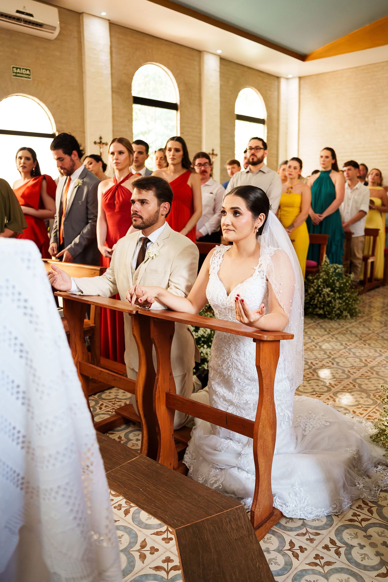 Bride and groom kneeling at church altar during wedding ceremony with guests standing in background.