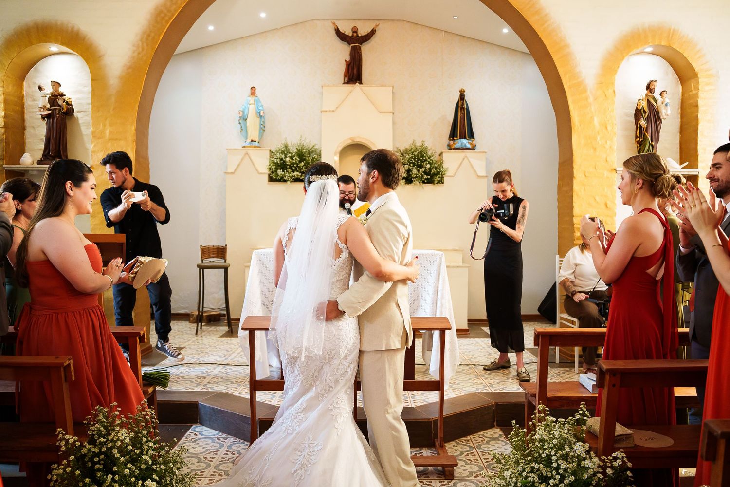 Bride and groom share first kiss at church altar surrounded by applauding guests and bridesmaids in red dresses.