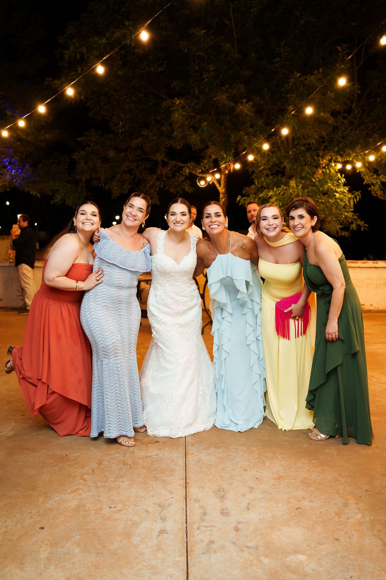 Bride in white gown poses with five bridesmaids in colorful dresses under string lights at outdoor wedding reception.