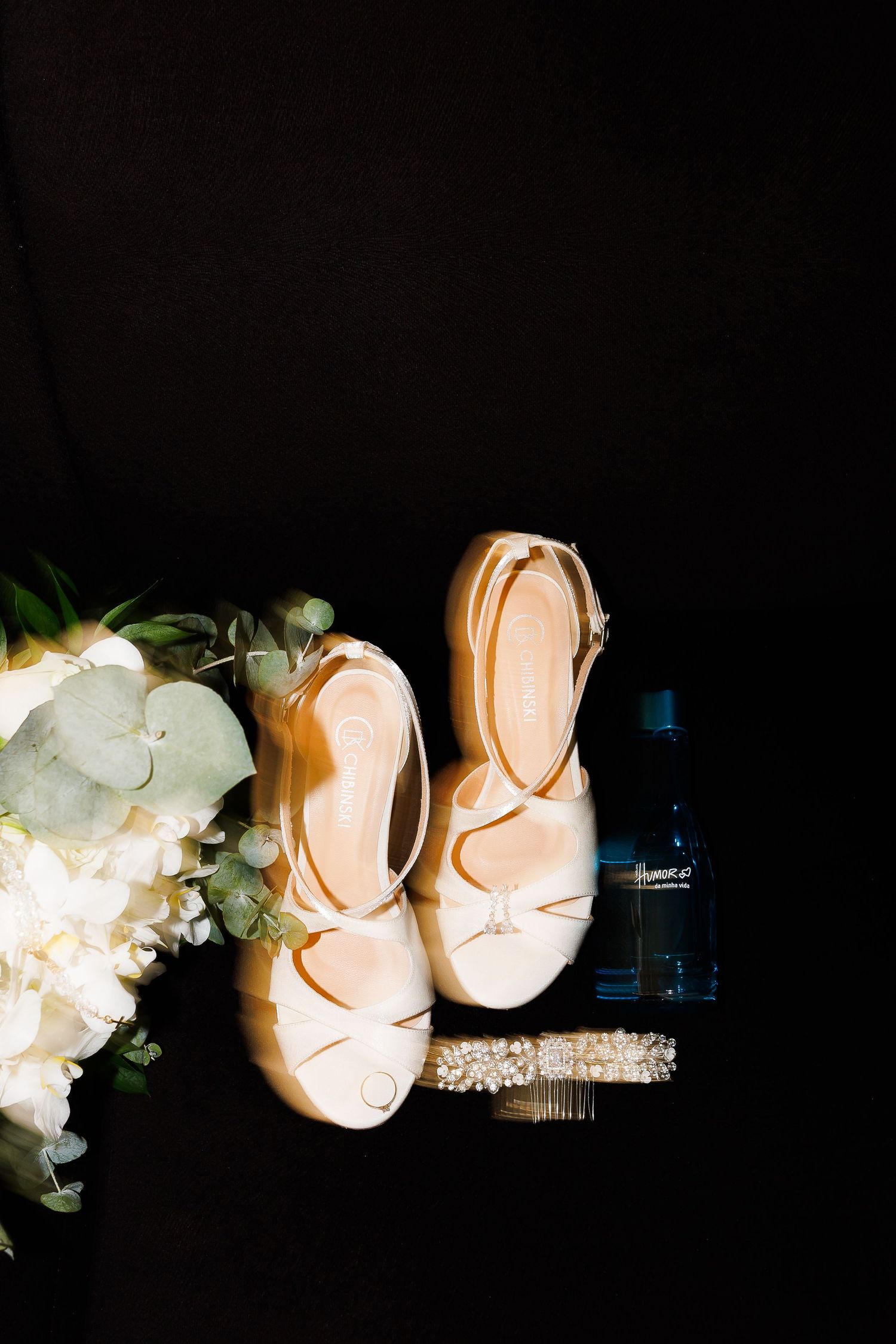 Elegant bridal heels with strappy design displayed beside a white floral bouquet and perfume bottle on black background.