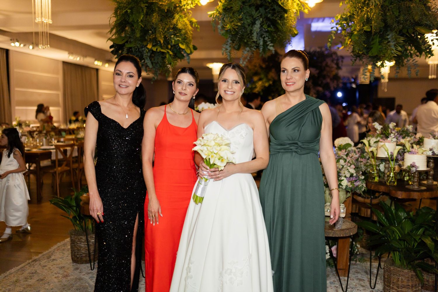 Bride in white gown holding bouquet poses with three elegantly dressed women at a wedding reception.