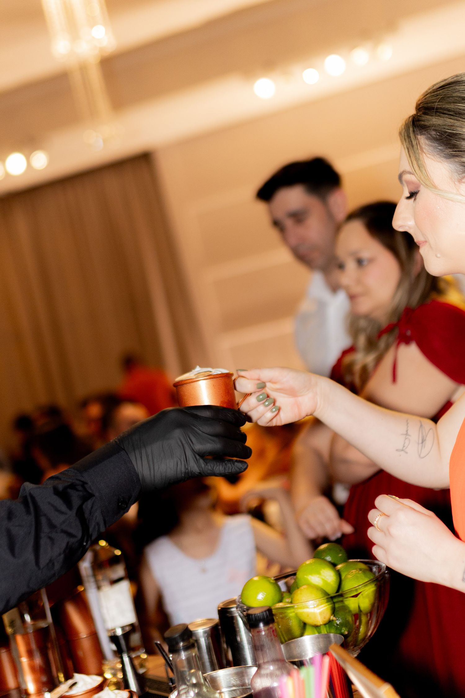 Bartender in black gloves hands a copper mug cocktail to a guest at an elegant event bar with limes.