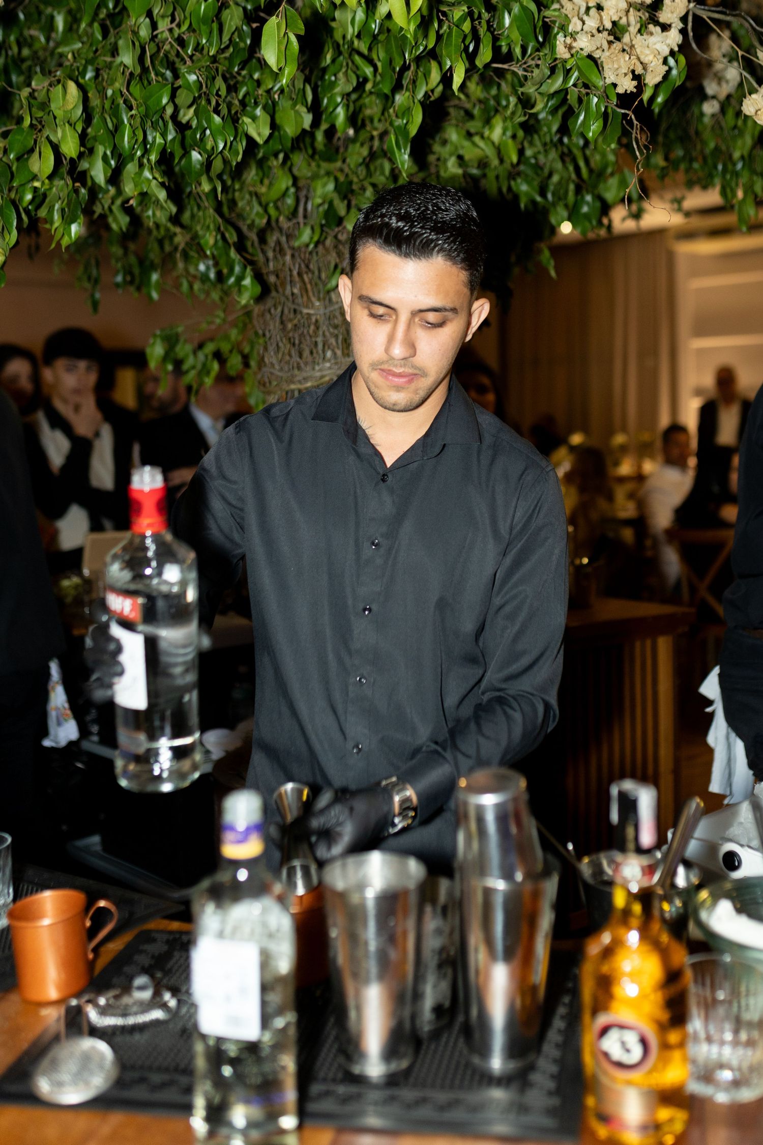 A bartender in black attire prepares cocktails at a bar with shakers and bottles under lush green foliage.
