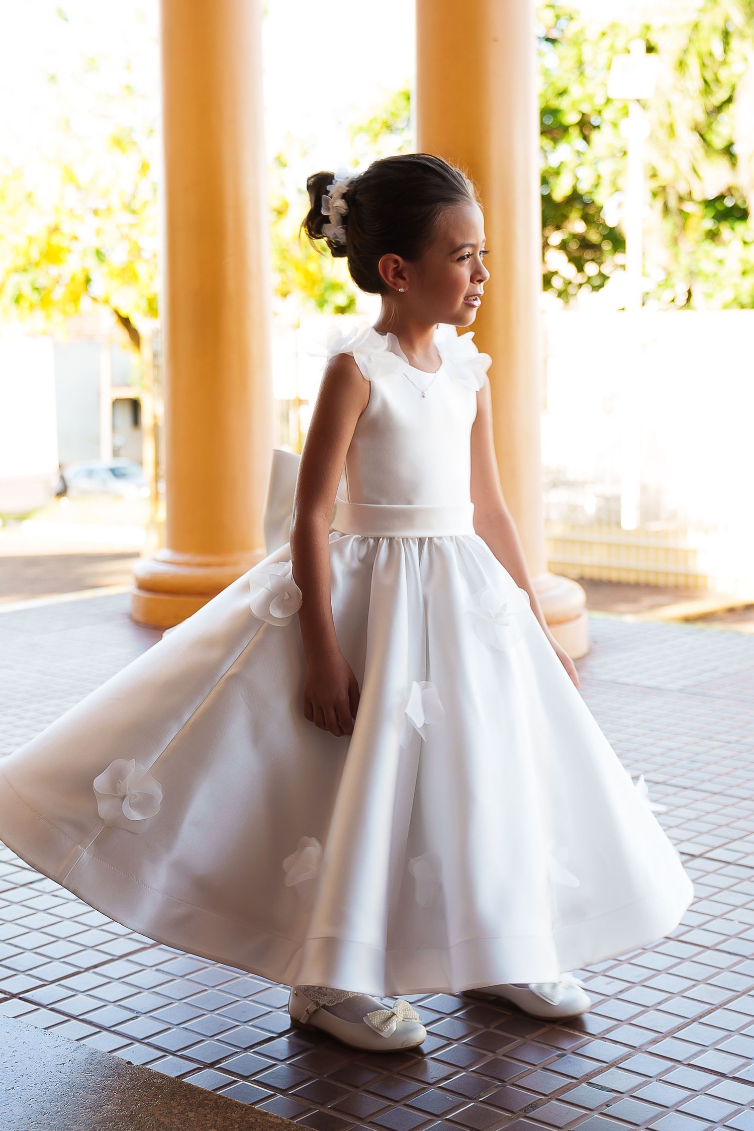 Young girl in white flower girl dress with floral details, posing between yellow columns outdoors.