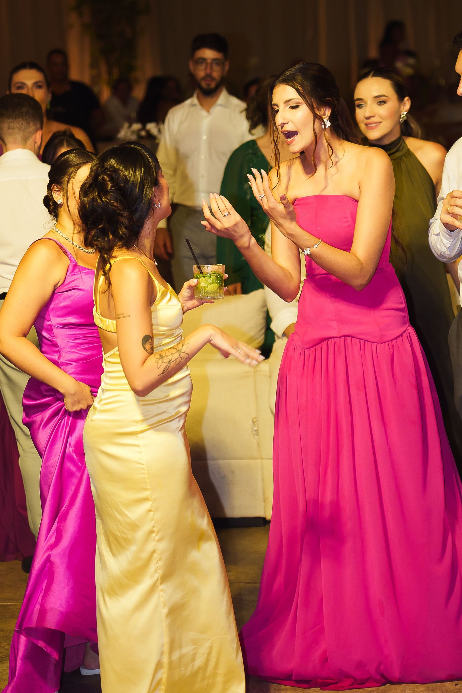 Women in colorful formal gowns dancing joyfully at an elegant indoor wedding reception celebration.