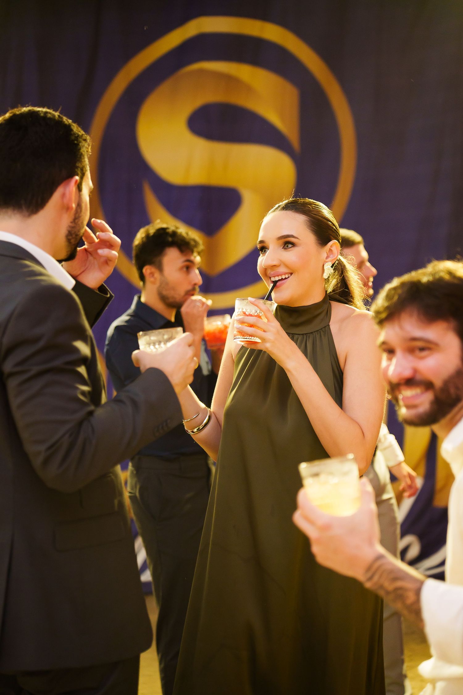 Elegantly dressed guests socializing at an upscale party, sipping cocktails in front of a gold and purple logo backdrop.