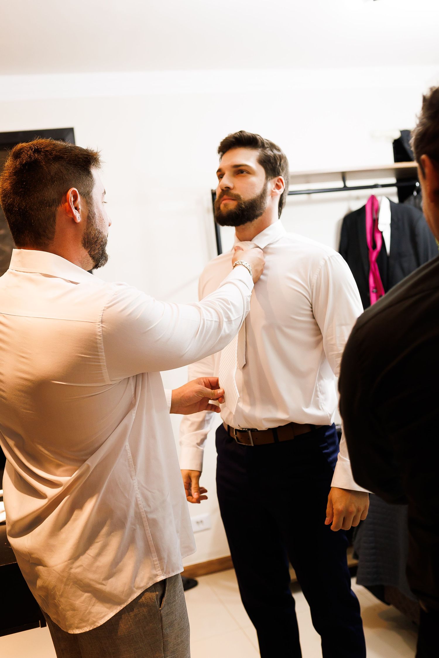 Man helping another man adjust his tie while getting dressed in formal wear.