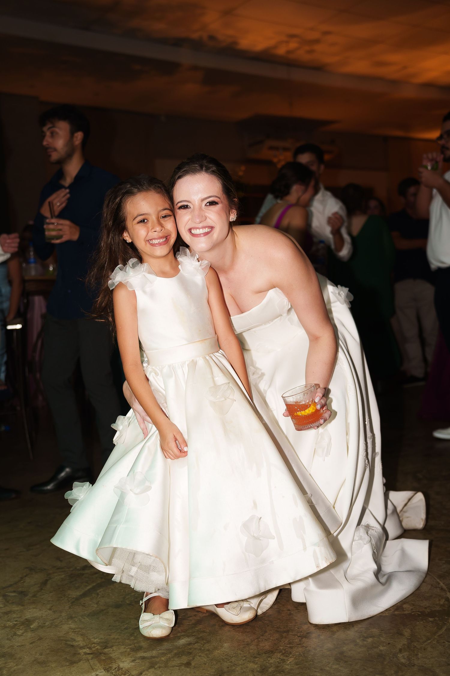Bride in white gown crouching beside flower girl in matching white dress, smiling at wedding reception.