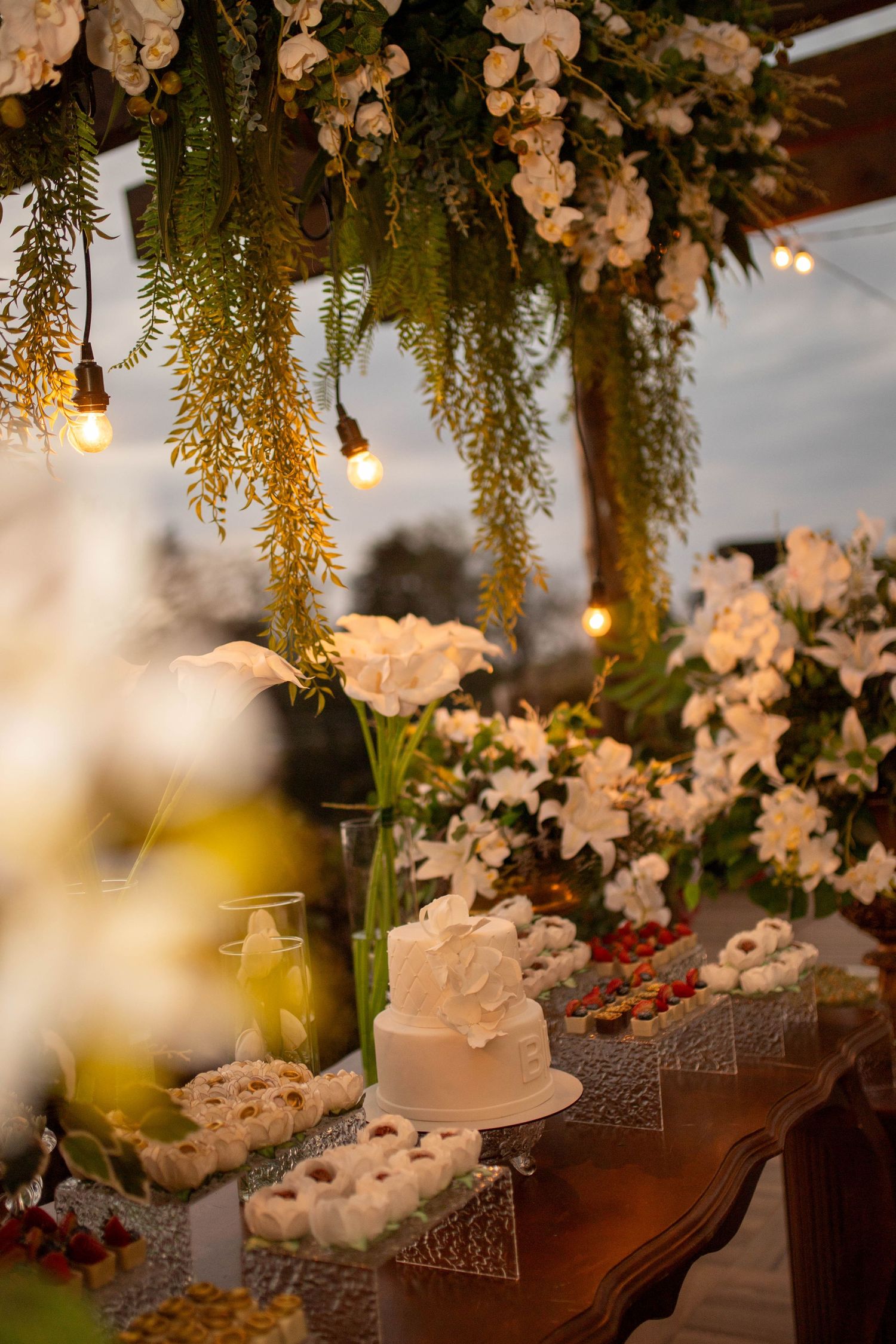 Elegant wedding dessert table with white cake, flowers, string lights, and lush floral arch overhead.