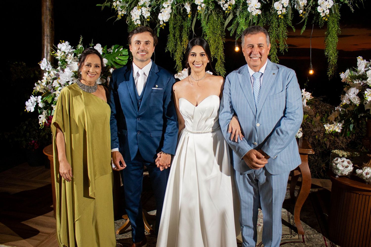 Wedding party of four posing outdoors at night, bride in white gown, groom in blue suit, flanked by two guests.
