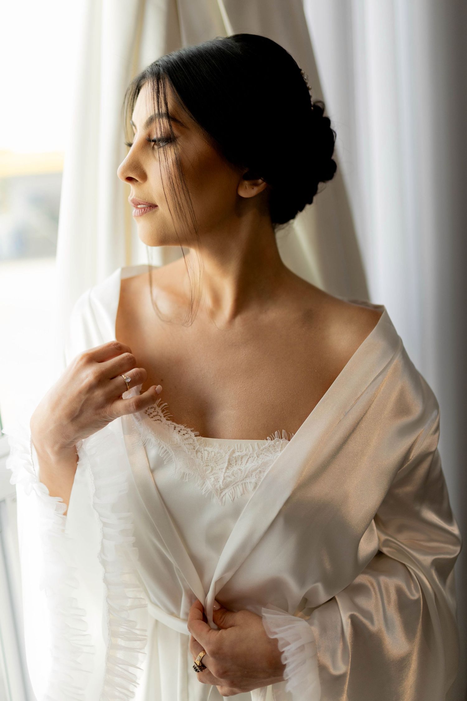 Elegant bride in white satin robe gazing out window with updo hairstyle and ring on wedding morning.