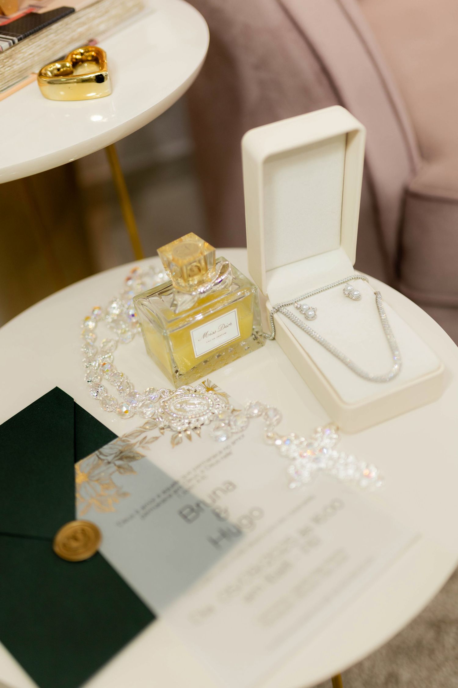 Elegant wedding flat lay with Miss Dior perfume, diamond necklace in box, crystal bracelet, and green envelope invitation.