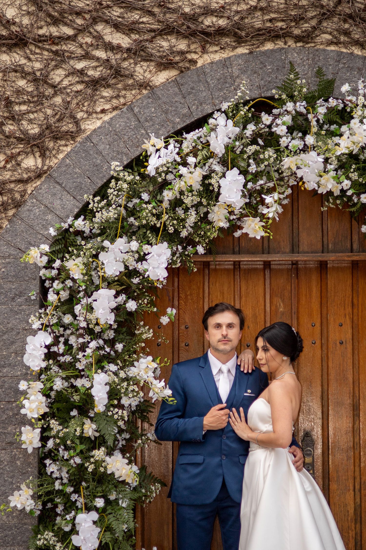 Bride and groom pose before a wooden door framed by a white floral arch at their elegant wedding ceremony.