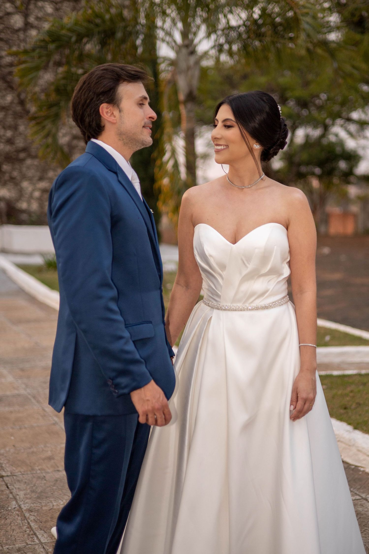 A bride in a white strapless gown and groom in a blue suit hold hands outdoors on their wedding day.