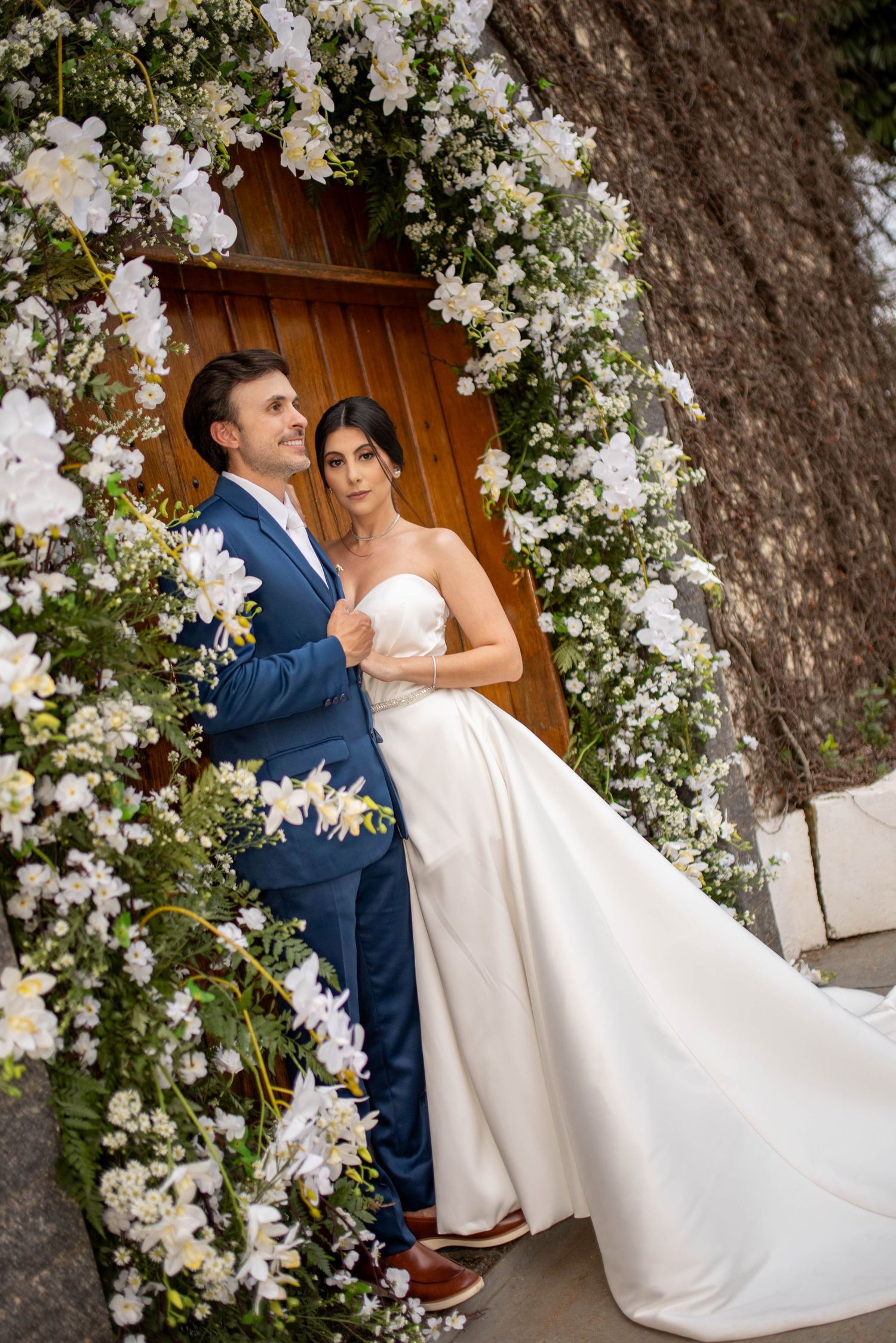 Bride and groom posing under a white floral arch with a wooden door backdrop on their wedding day.