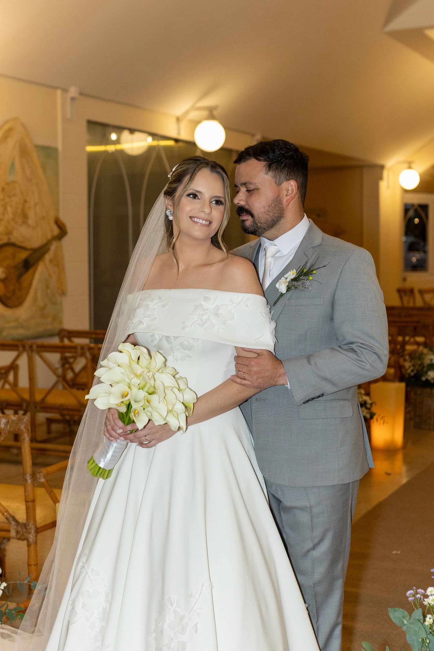 Bride in white off-shoulder gown holding calla lily bouquet poses with groom in gray suit inside a church.