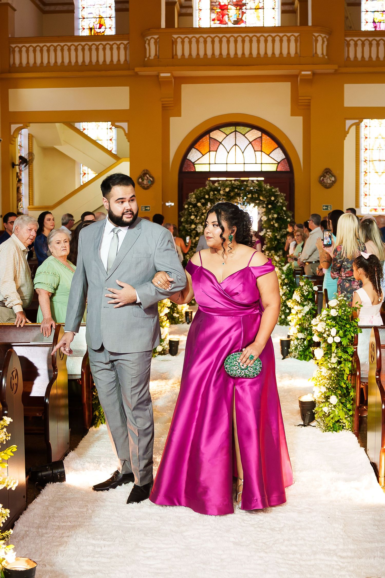 A man in a gray suit escorts a woman in a magenta satin gown down a decorated church aisle at a wedding.