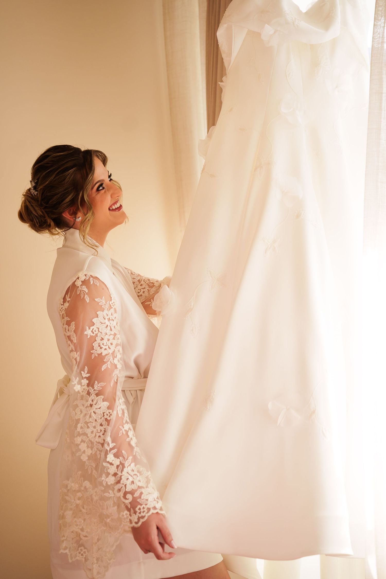 Smiling bride in lace robe admiring her white wedding dress hanging by a bright window.