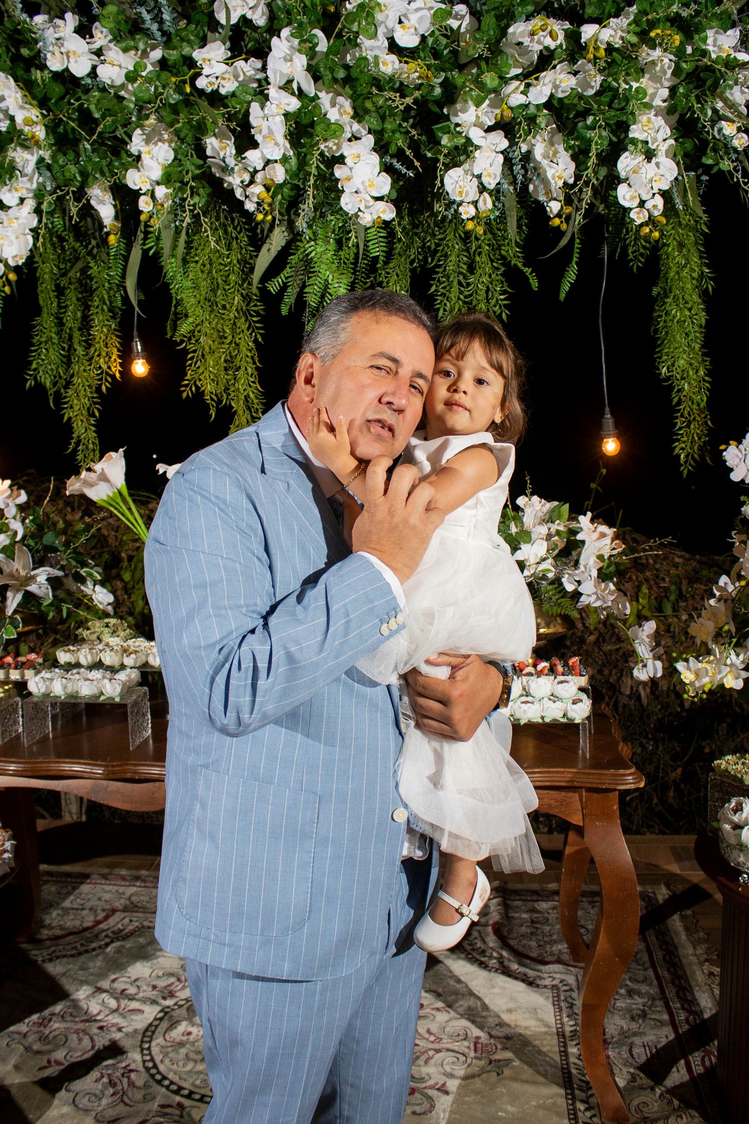 Man in blue suit holding a young girl in white dress at a floral decorated event.