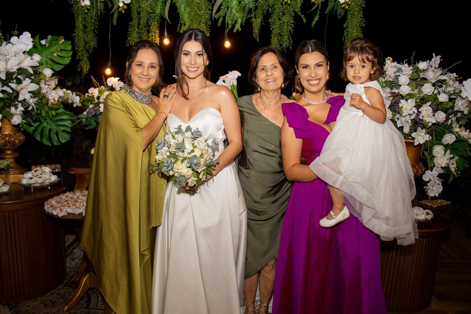 Bride in white gown poses with four women including bridesmaids and flower girl at elegant floral wedding reception.