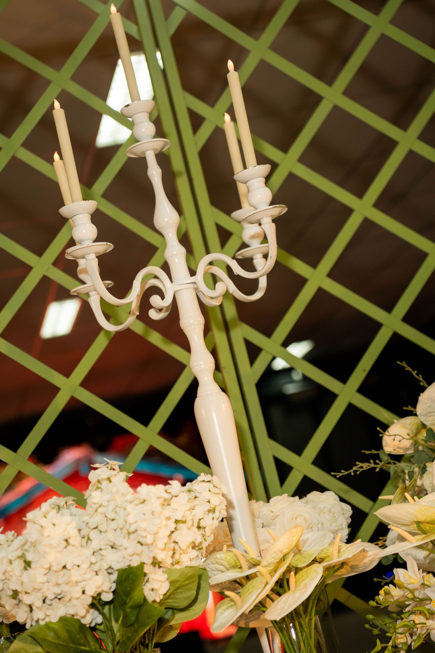 White candelabra with unlit candles surrounded by white hydrangeas and floral arrangements against a green lattice backdrop.
