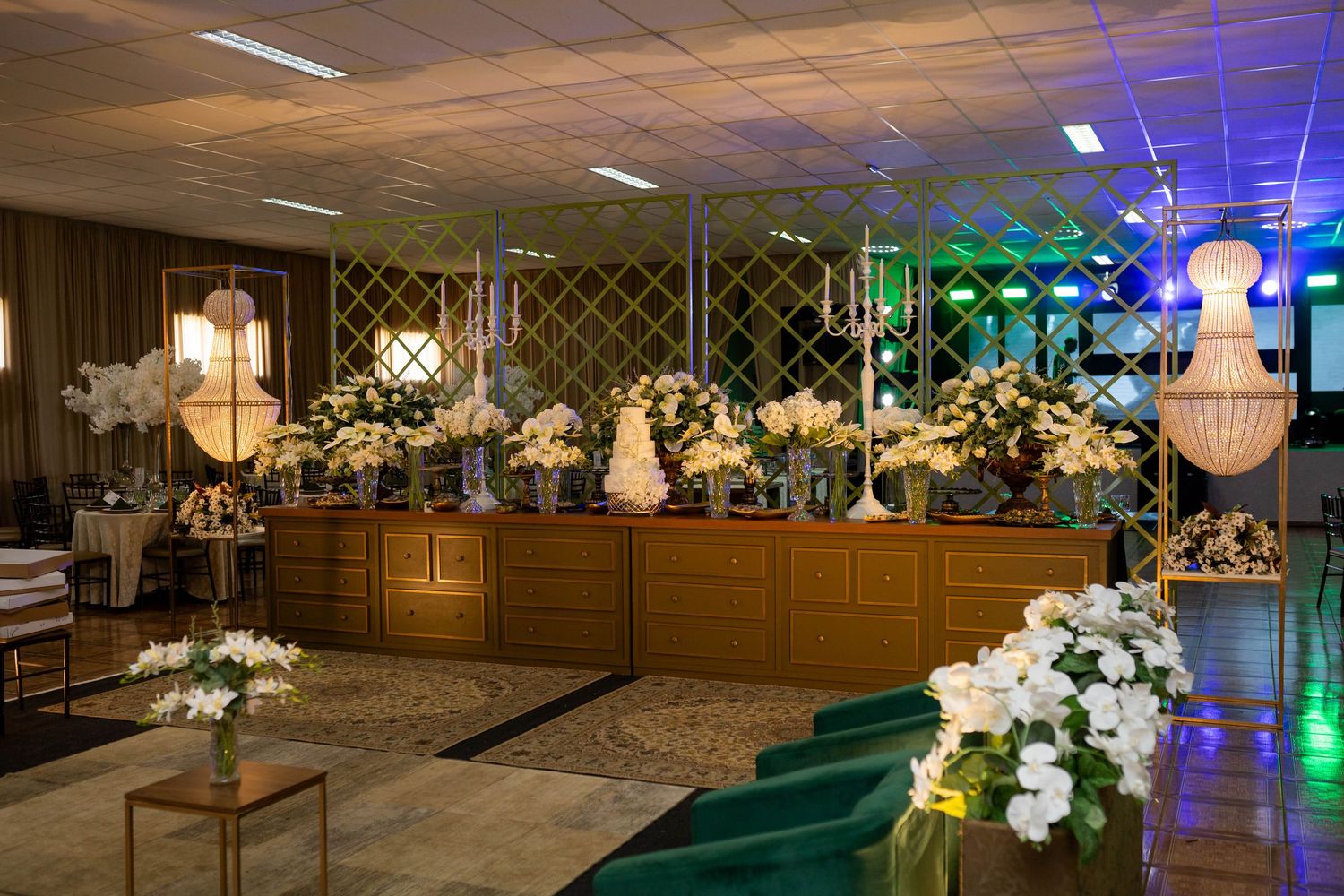 Elegant event hall with gold buffet table, white floral arrangements, green velvet chairs, and chandelier lighting.