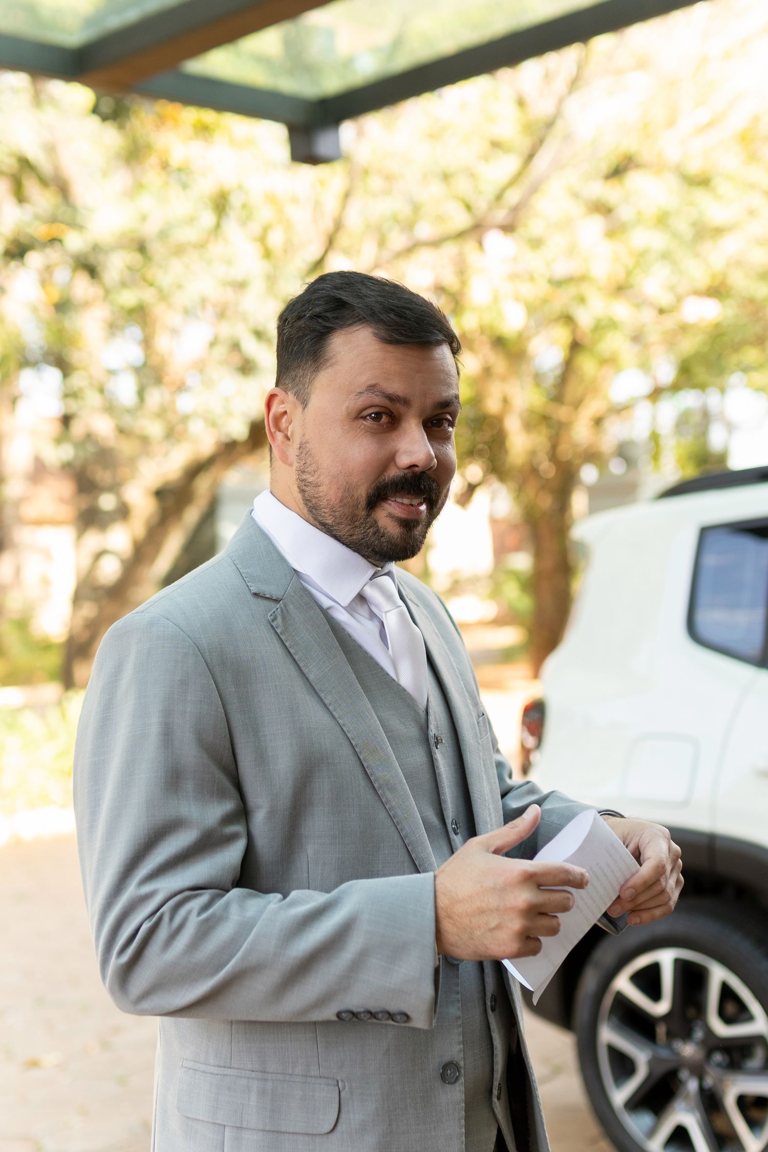Man in gray suit holding documents outdoors near white SUV, smiling confidently in natural setting.