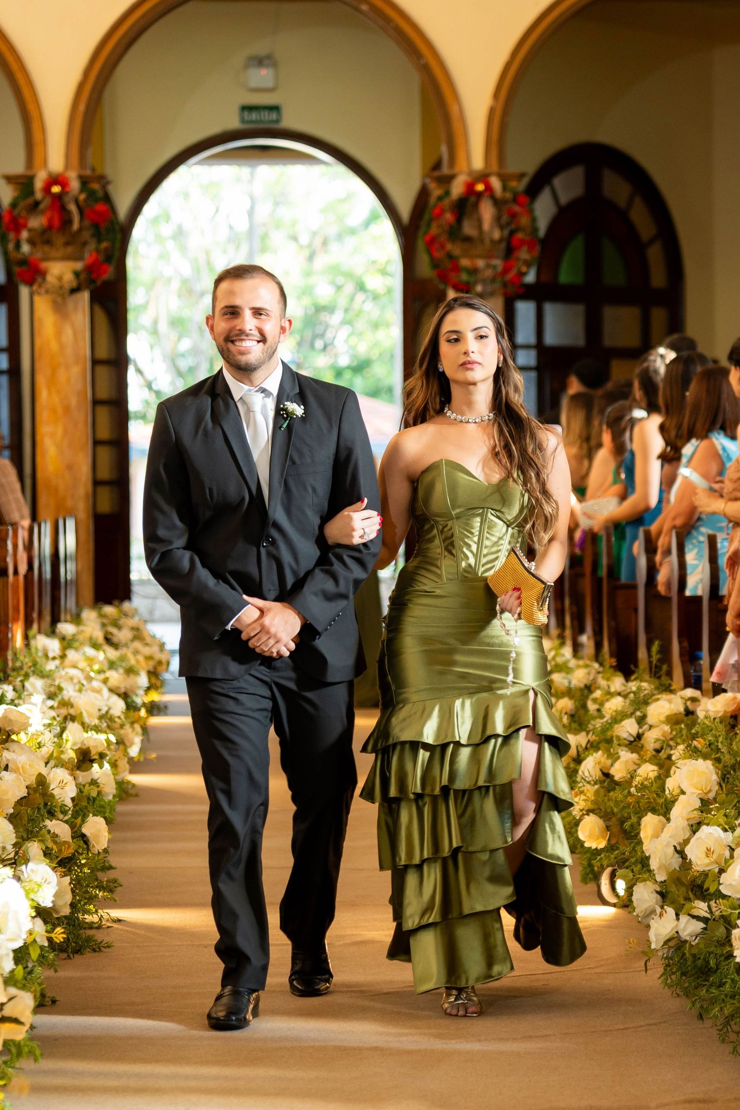 Groomsman in black suit walks bridesmaid in olive green ruffled dress down floral wedding aisle.