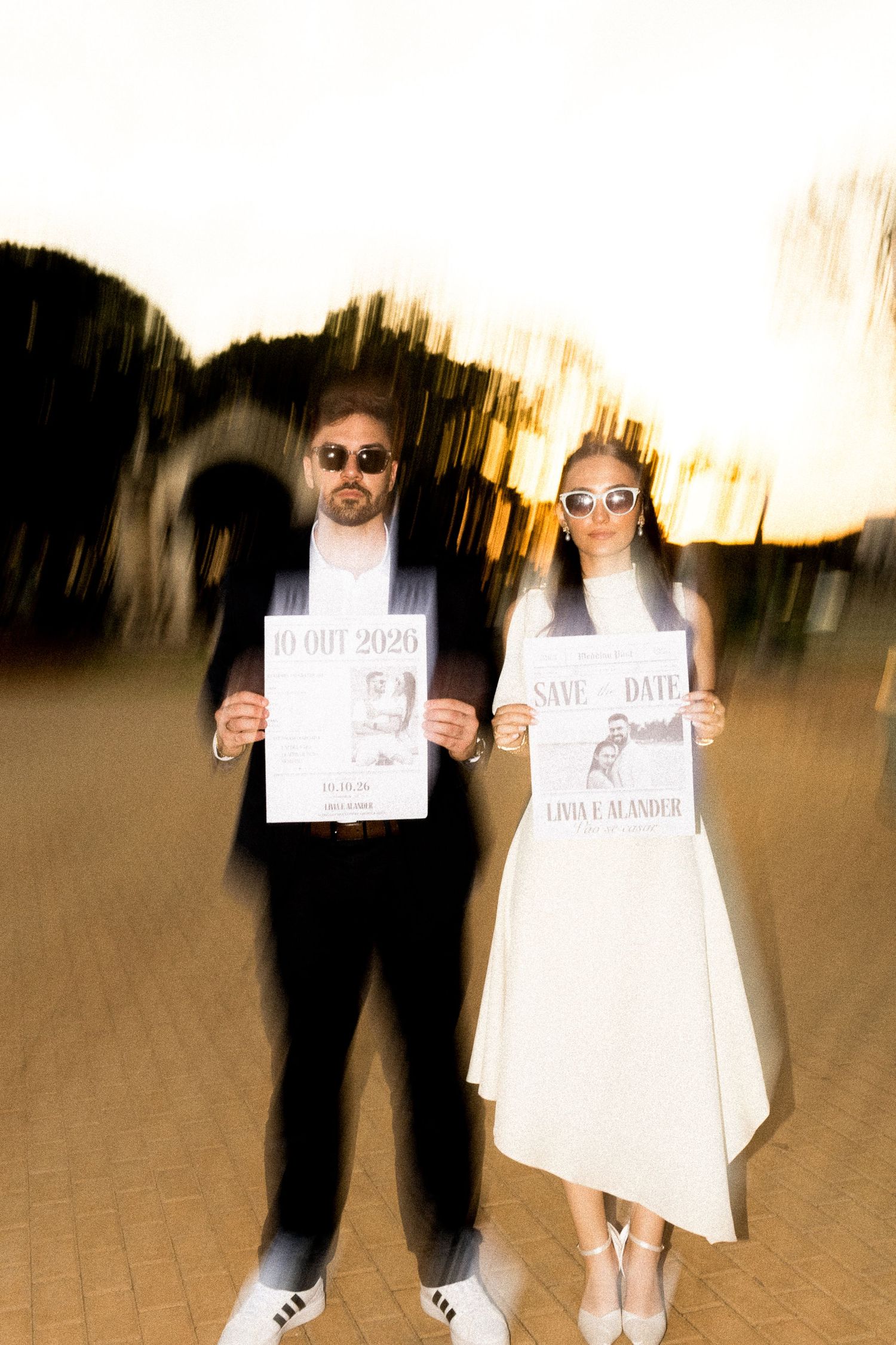 Couple in sunglasses holding Save the Date wedding signs outdoors with artistic motion blur effect.