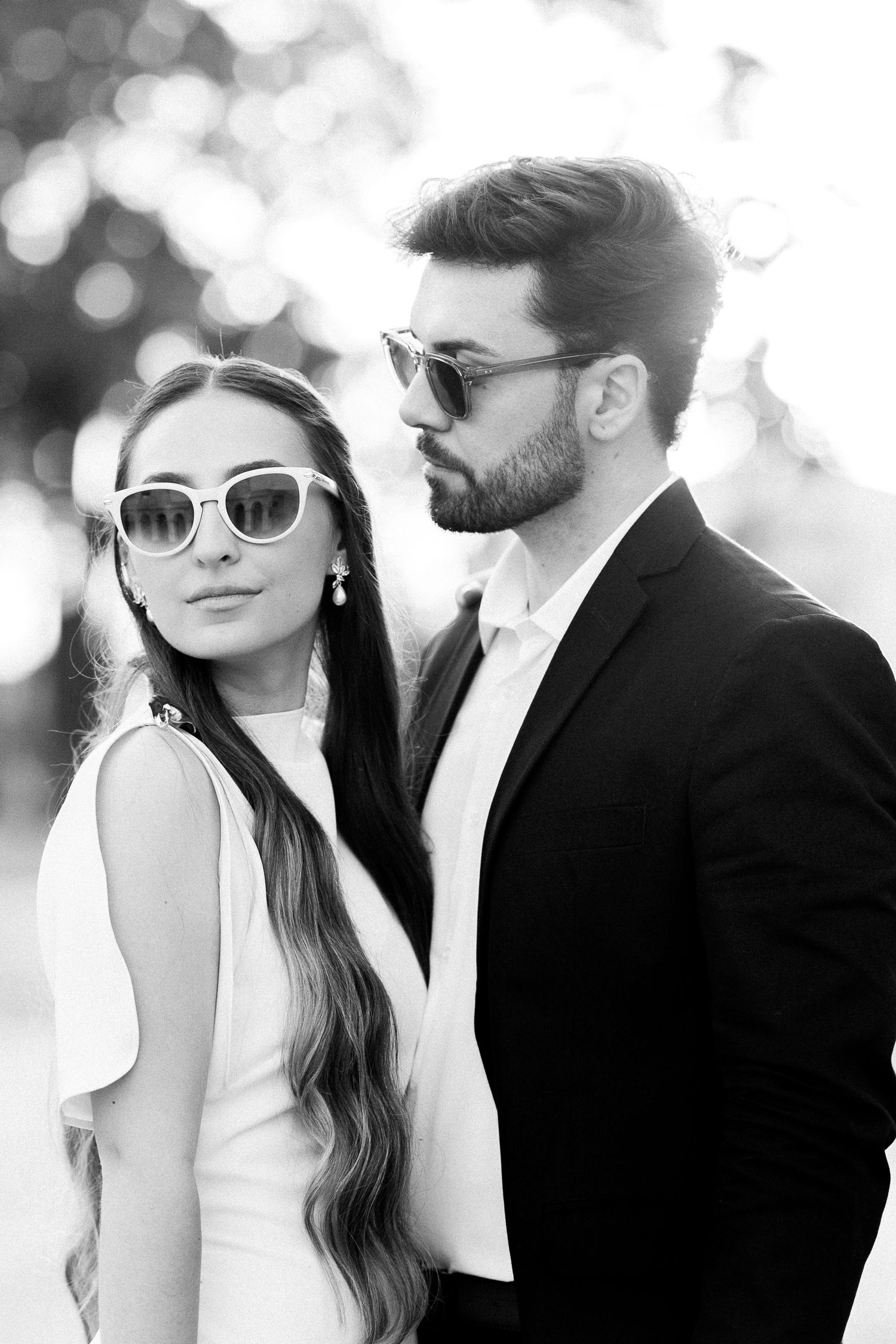 Black and white photo of a stylish couple wearing sunglasses, man in suit and woman in white dress outdoors.