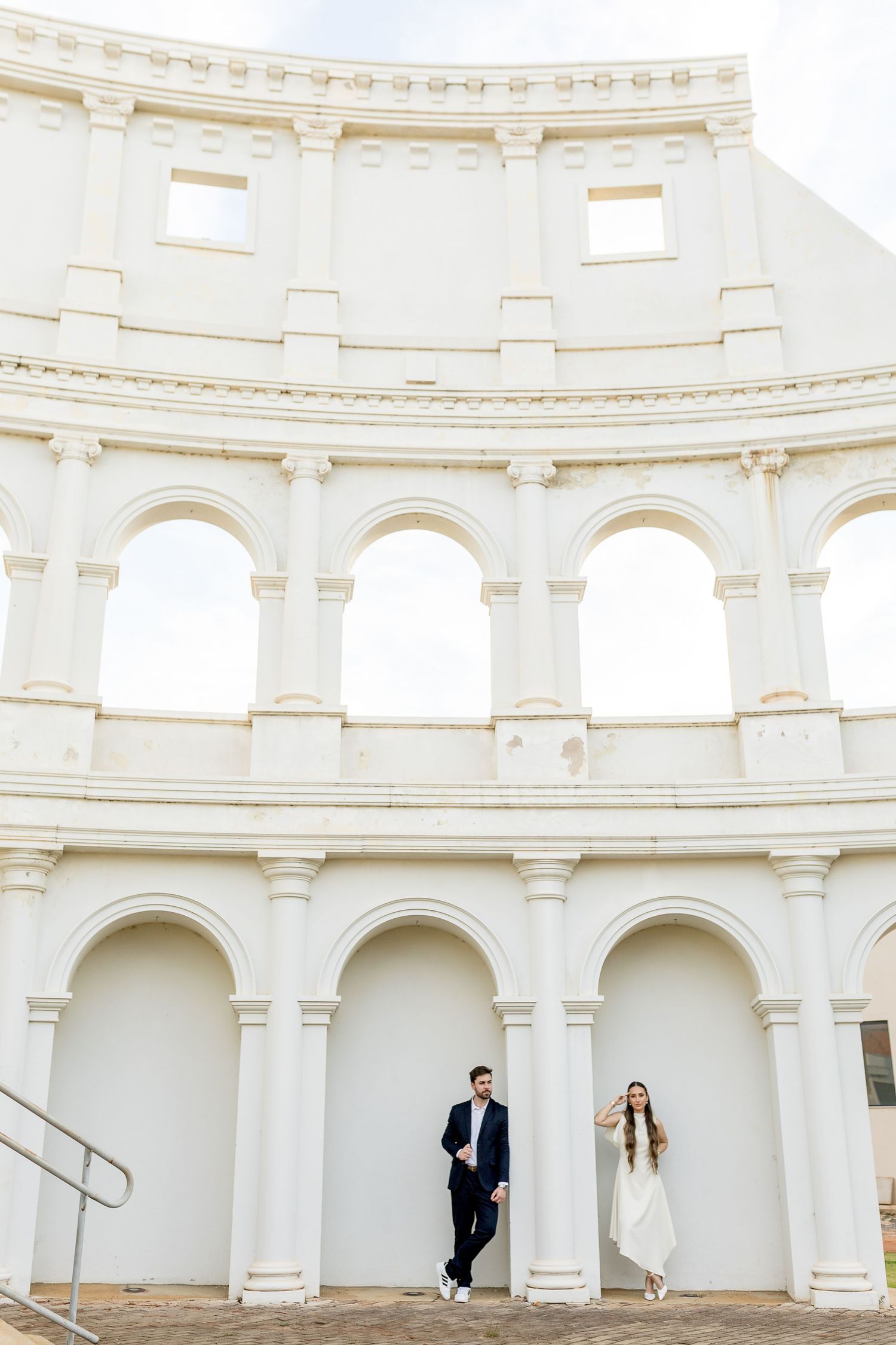 Couple posing in front of a white neoclassical building with arched columns, reminiscent of the Colosseum.