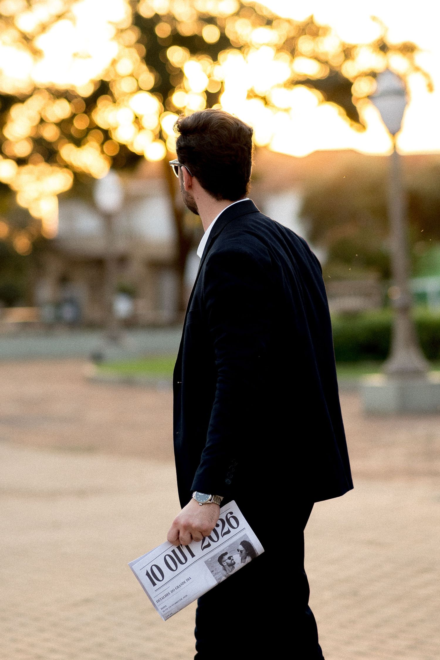 Man in dark suit holding Save the Date newspaper walking at sunset with golden bokeh trees.
