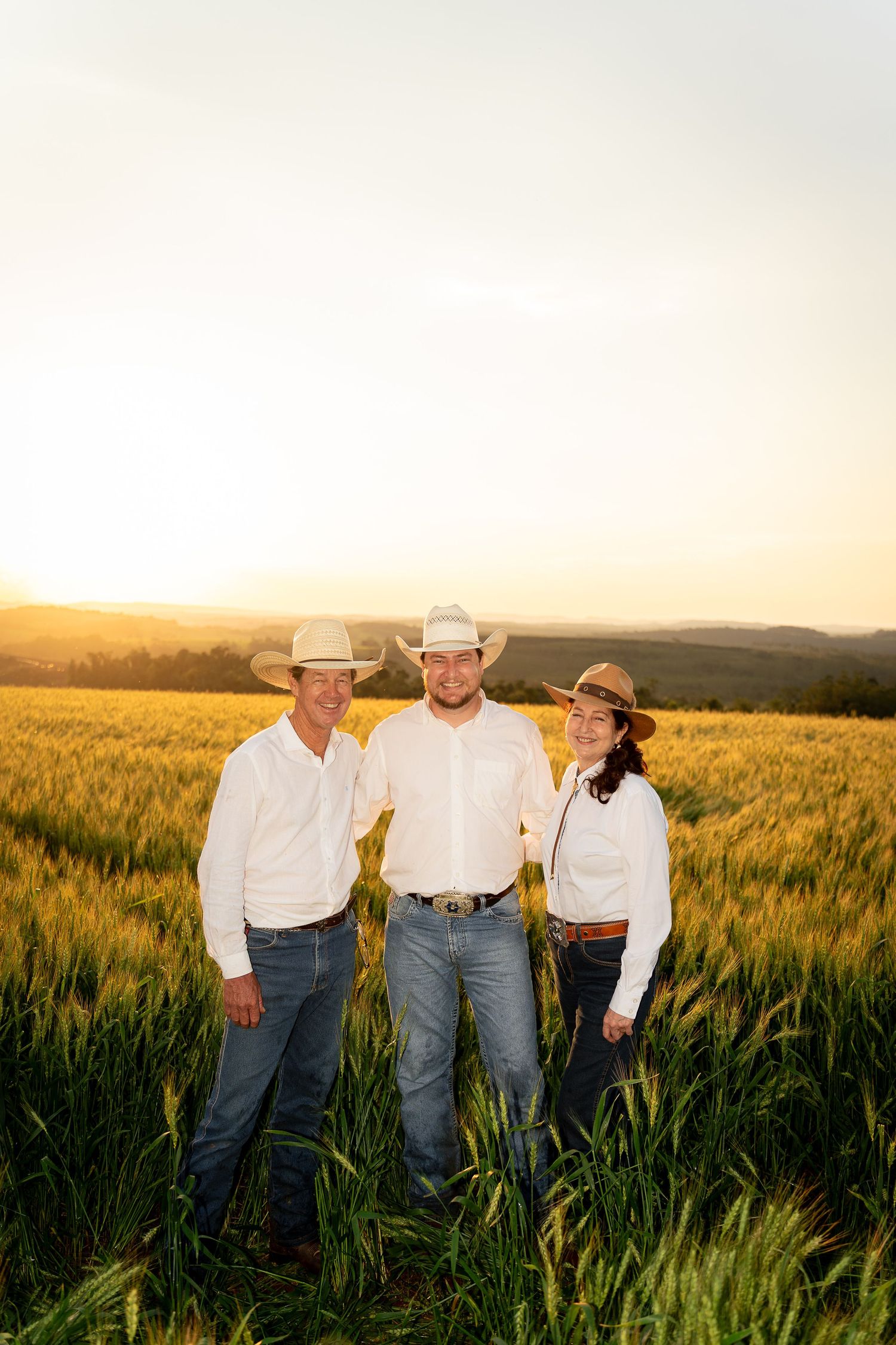 Three people in cowboy hats and white shirts stand smiling in a golden wheat field at sunset.