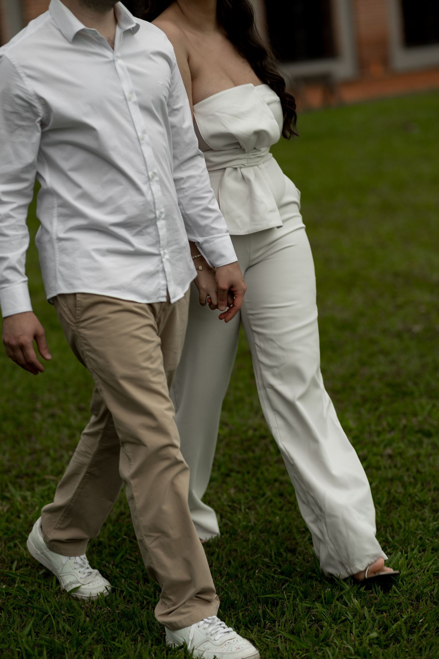 Couple holding hands walking on grass, man in white shirt and khakis, woman in white strapless top and wide-leg pants.