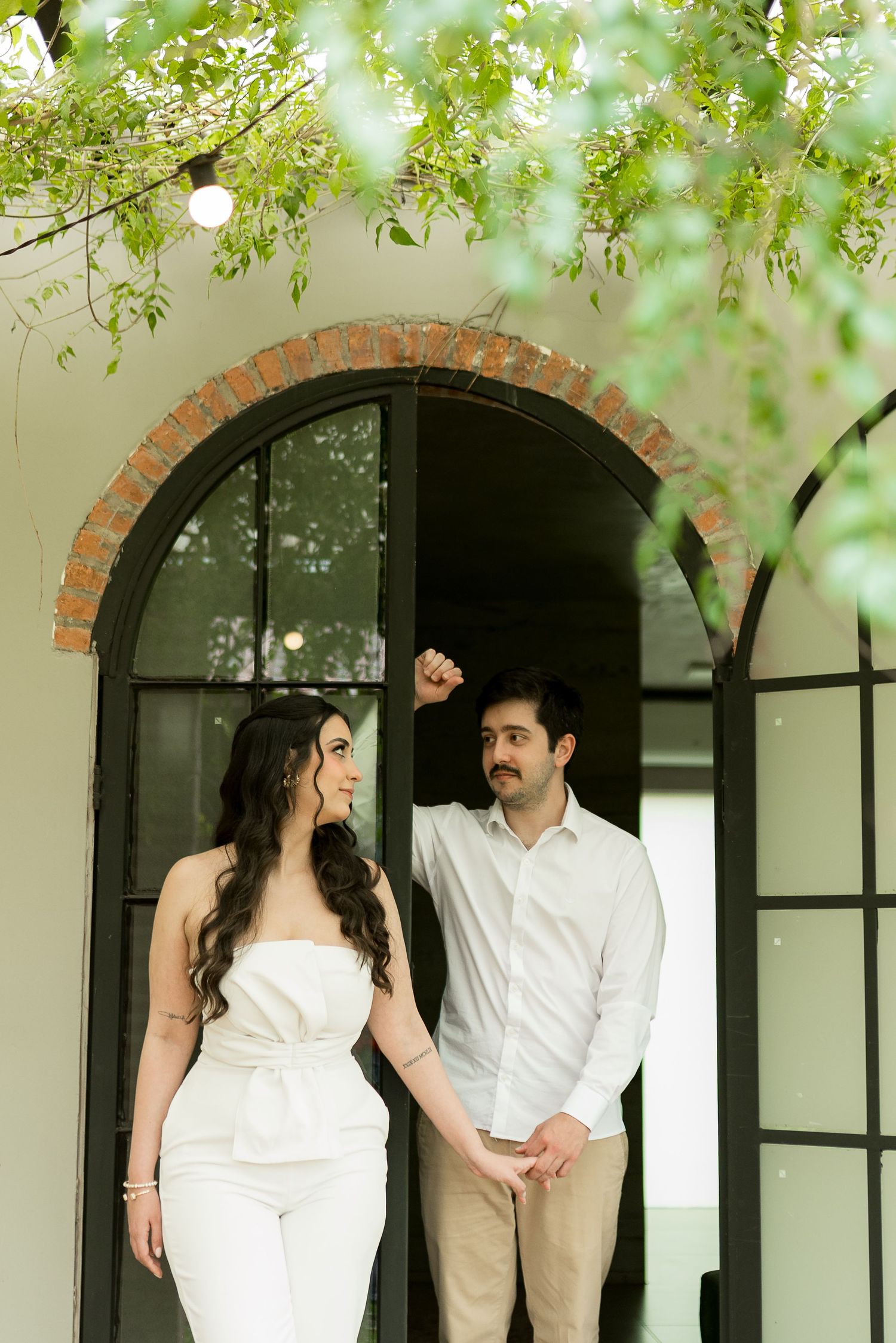 Couple holding hands and dancing under arched brick doorway surrounded by lush green trees in elegant white outfits.