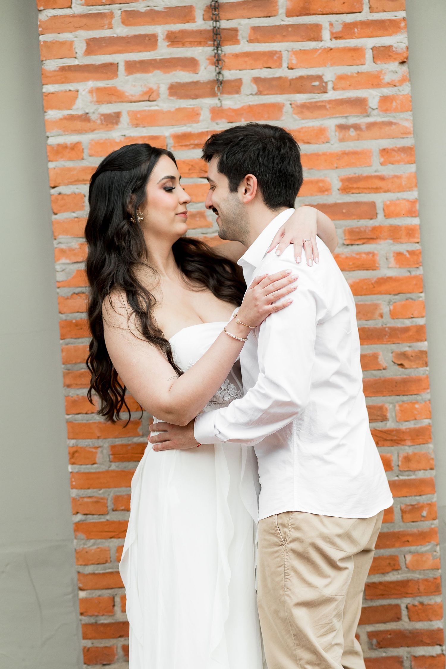 Romantic couple in white outfits embracing near a red brick wall, sharing a tender moment.
