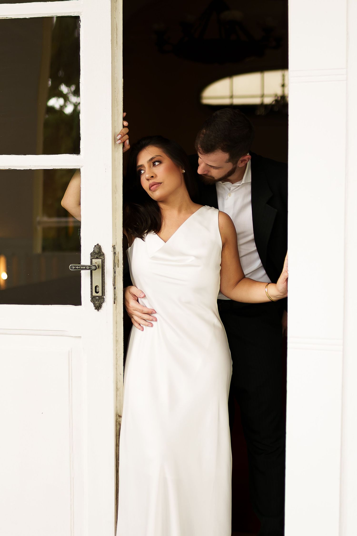 Bride in white gown stands in doorway as groom in tuxedo embraces her from behind at elegant wedding venue.
