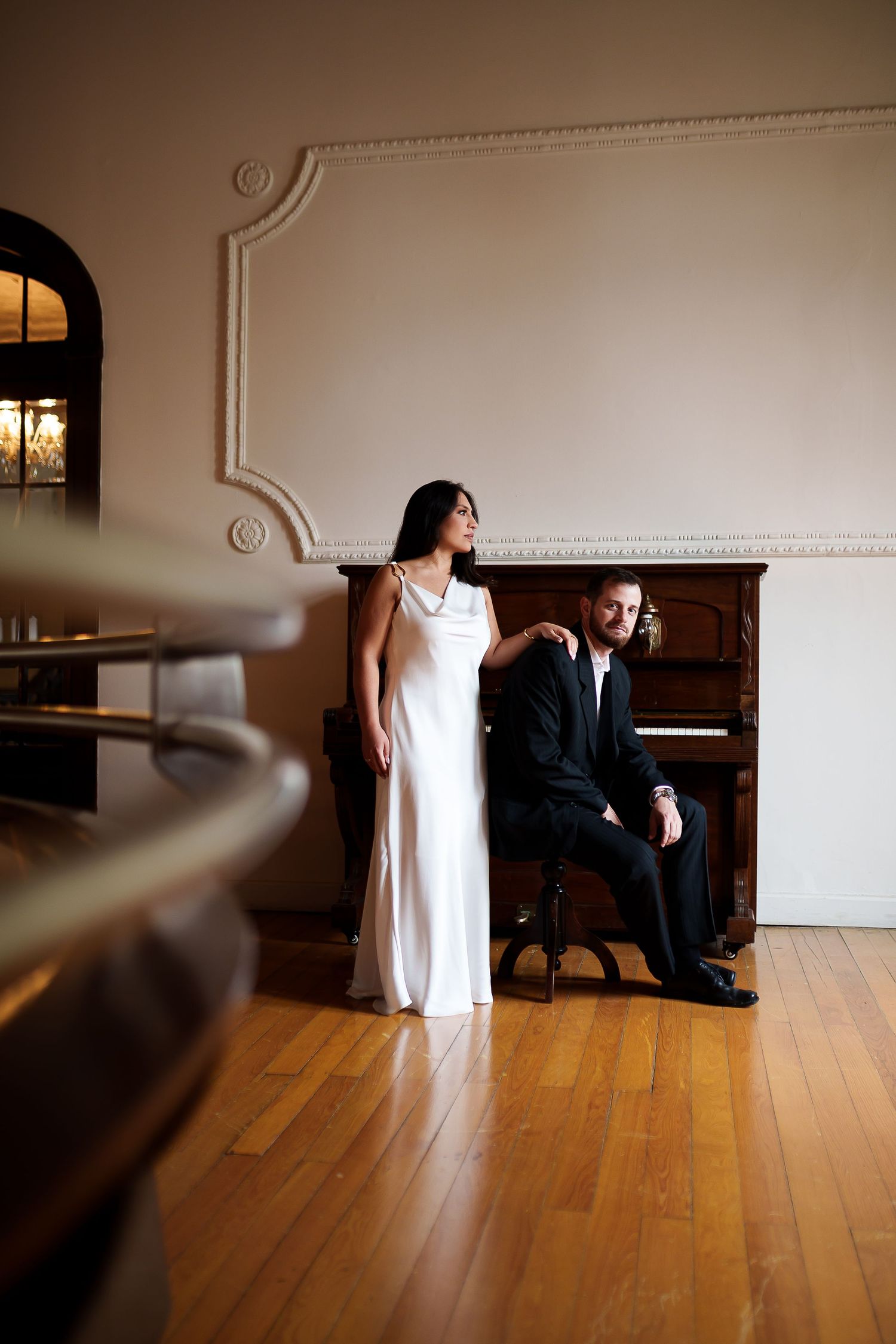 Elegant couple in formal attire posing by a grand piano in a classic white room with ornate moldings.