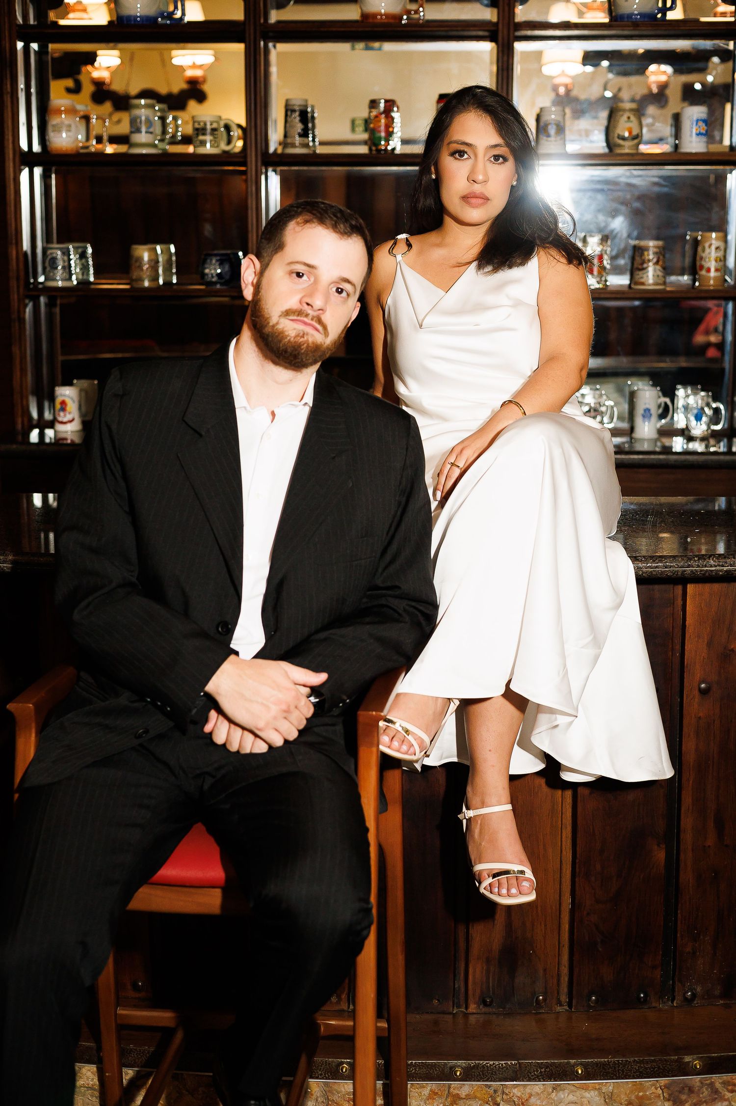 Couple in formal attire posing in a bar, man in black suit seated, woman in white dress perched on cabinet.