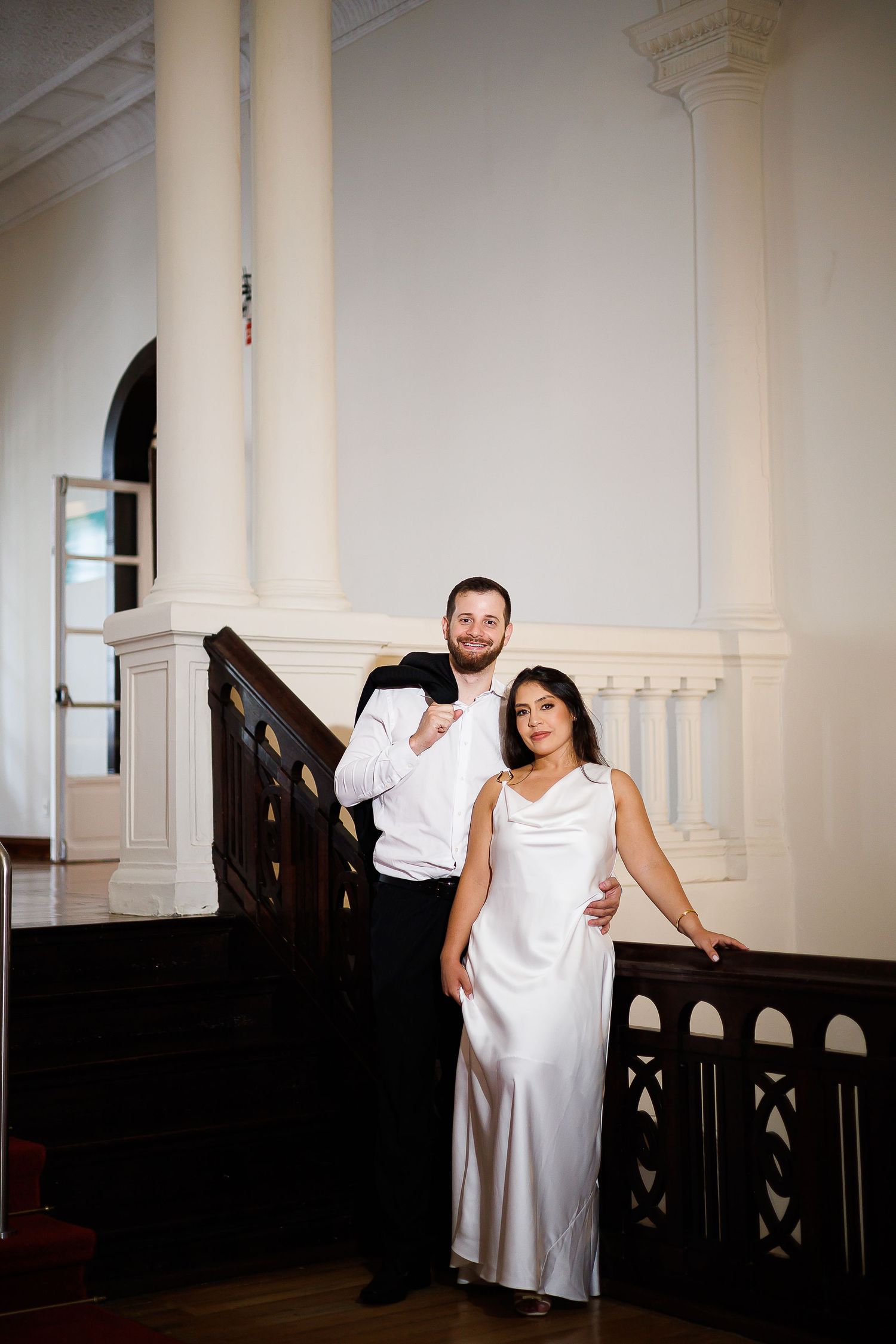 Elegant couple posing on grand staircase, woman in white satin gown, man in formal attire, classic white interior.