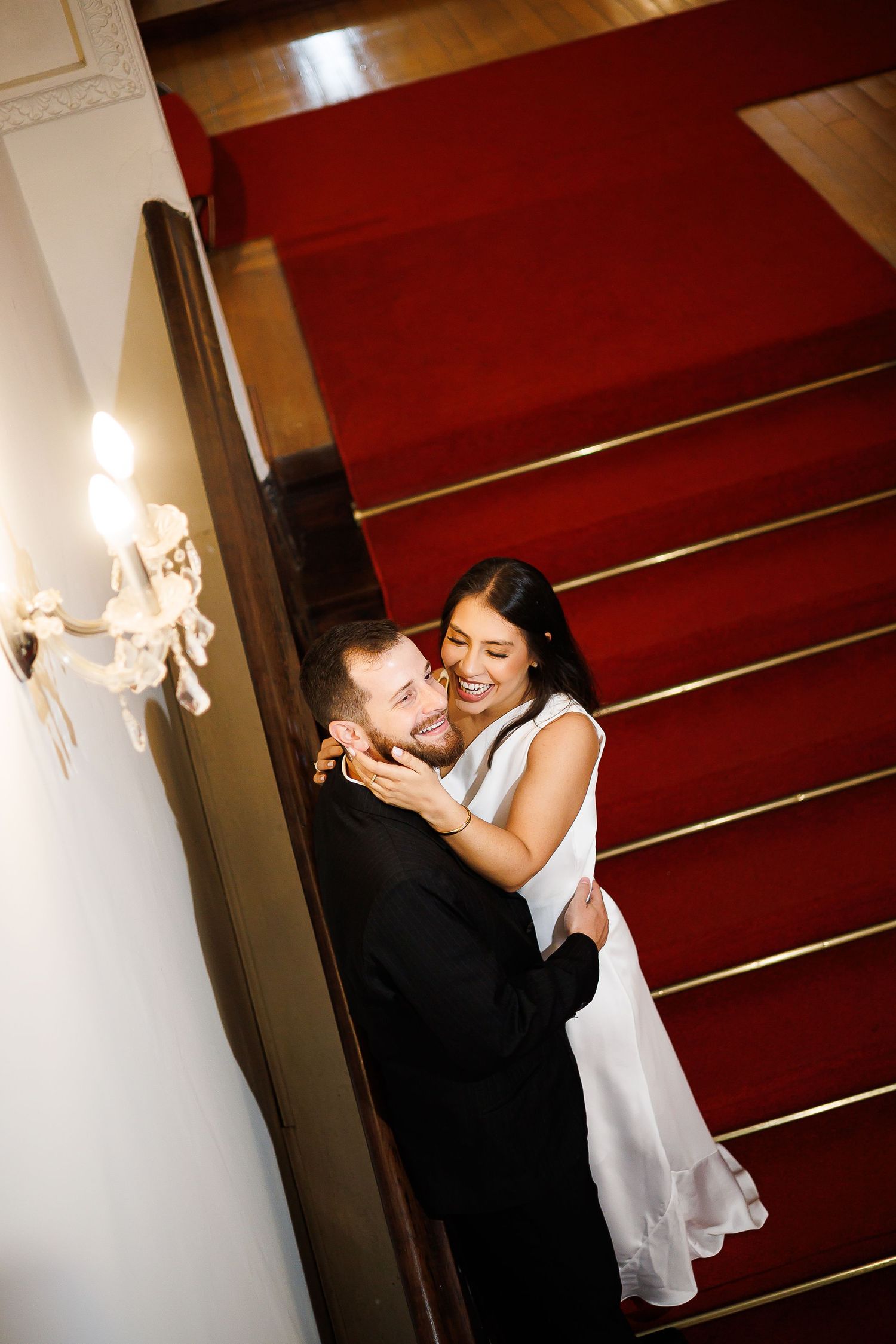 Bride and groom embracing and laughing on elegant staircase with red carpet runner in a grand venue.