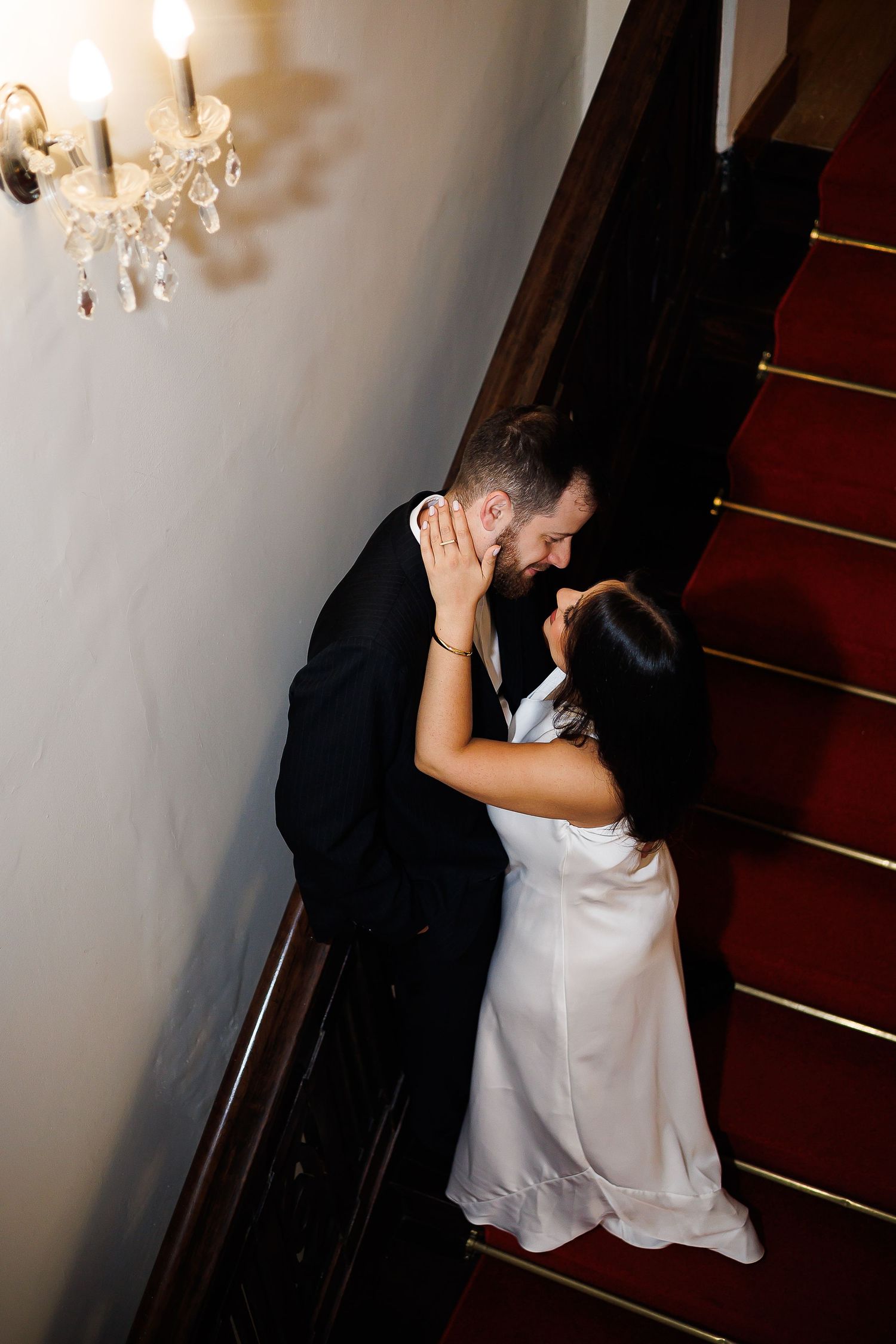Bride and groom share a romantic kiss on a grand staircase with red carpet and crystal chandelier.