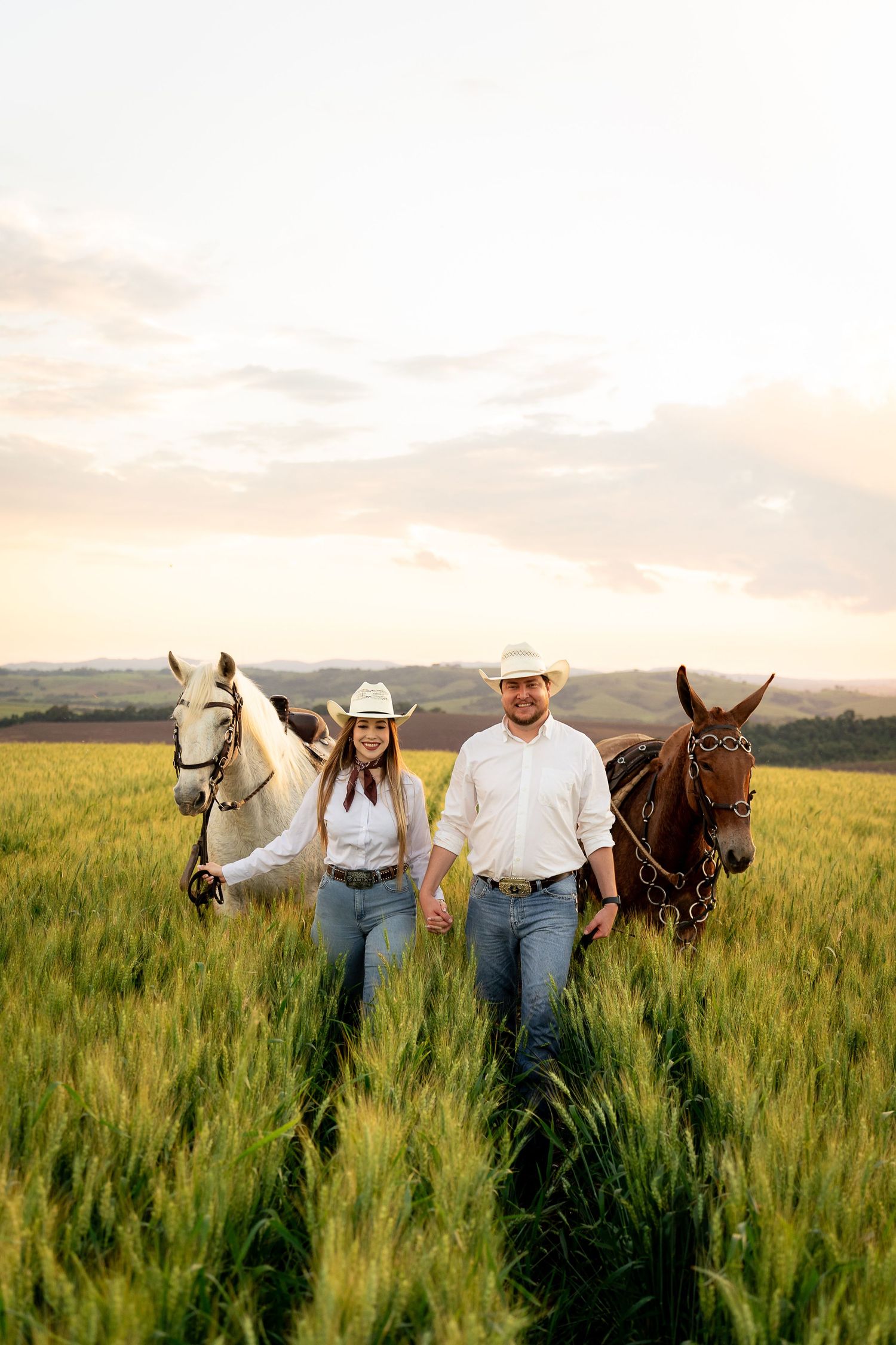 A smiling couple in cowboy hats walks hand-in-hand through a green field at sunset, leading a white and brown horse.