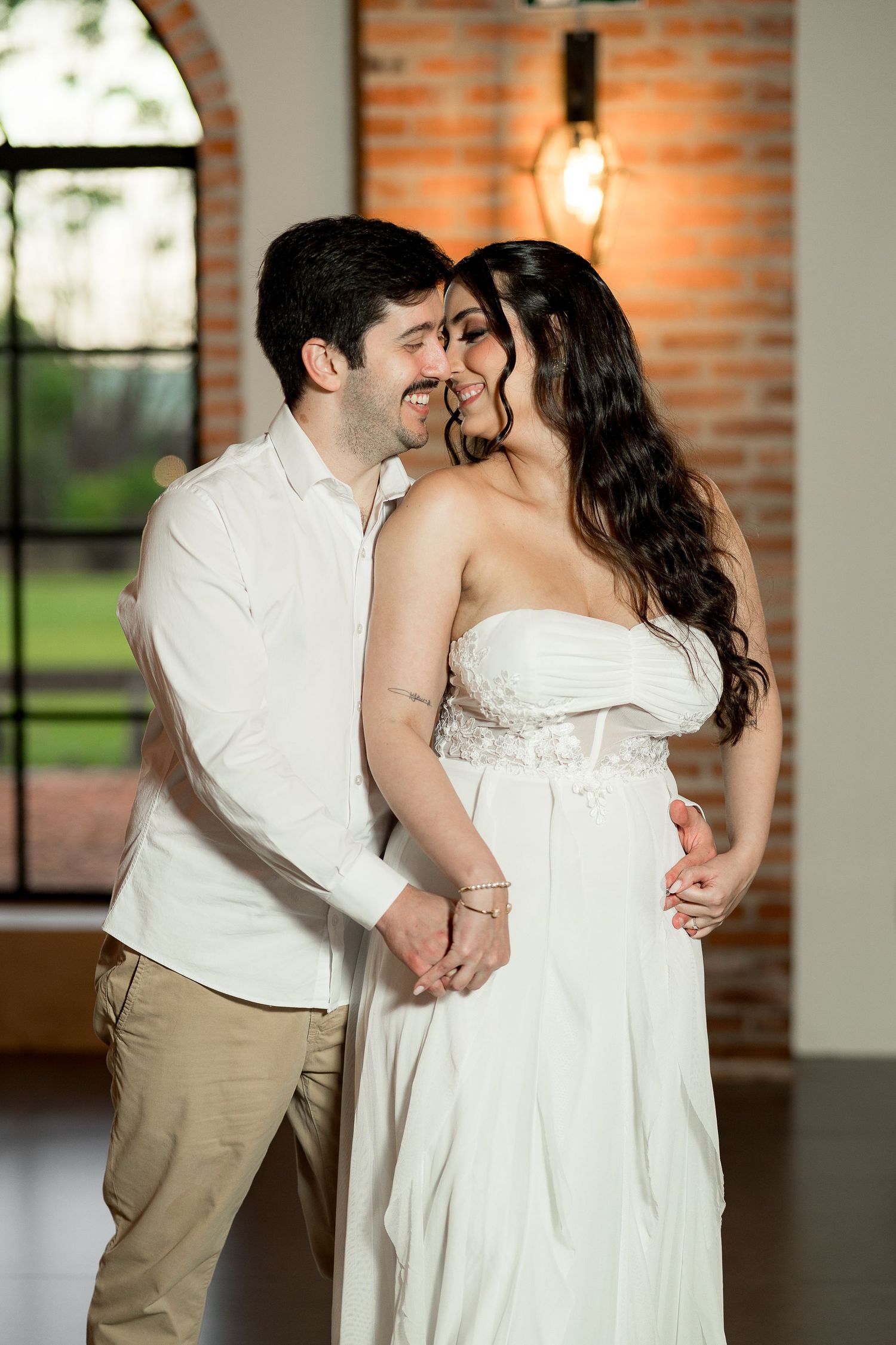A smiling couple in white outfits share a romantic dance in a rustic brick-walled venue with warm Edison bulb lighting.