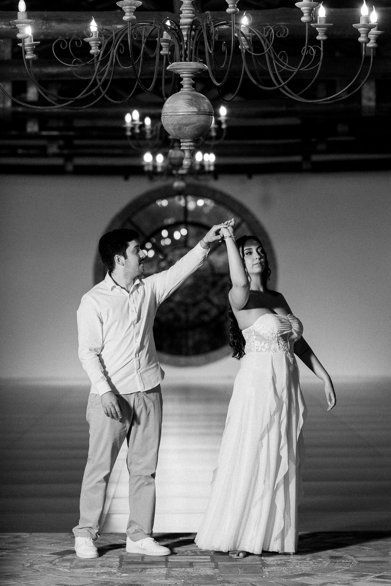 Black and white photo of a couple dancing elegantly under a chandelier in a grand hall, woman in white dress.