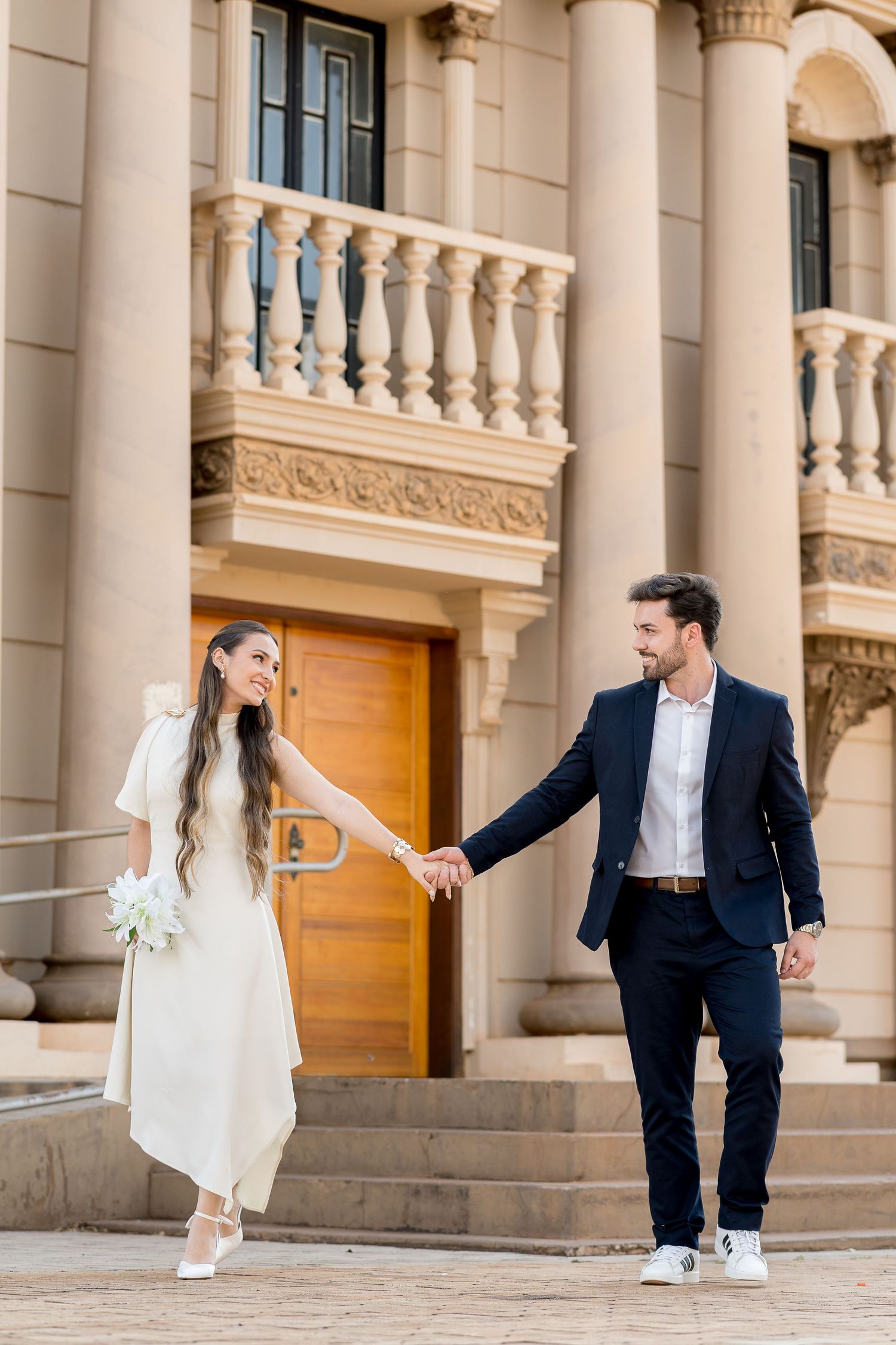 Bride and groom holding hands on steps of elegant stone building, bride in white dress holding bouquet, groom in navy suit.