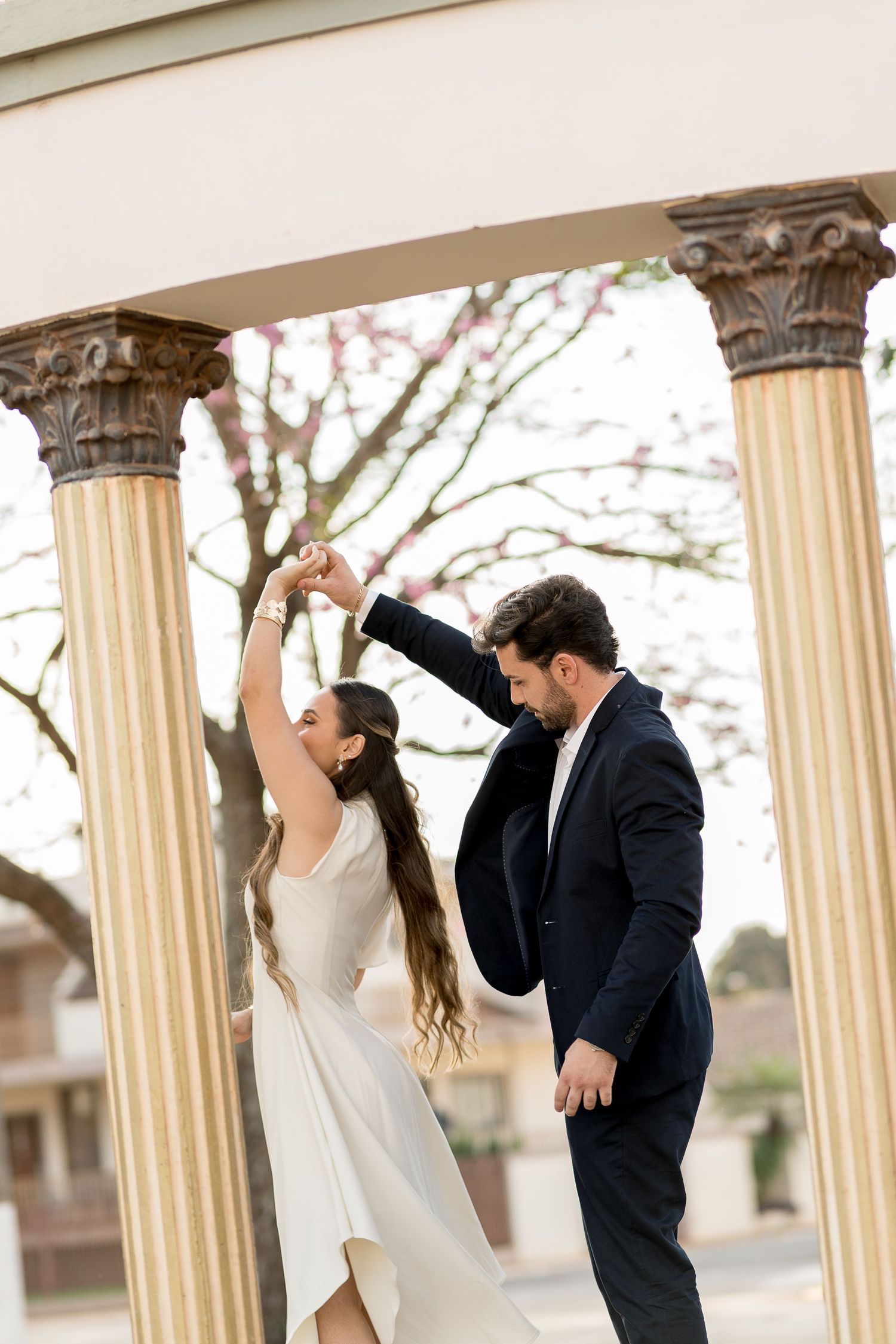Bride and groom dancing between elegant columns outdoors, with blooming trees in the background.