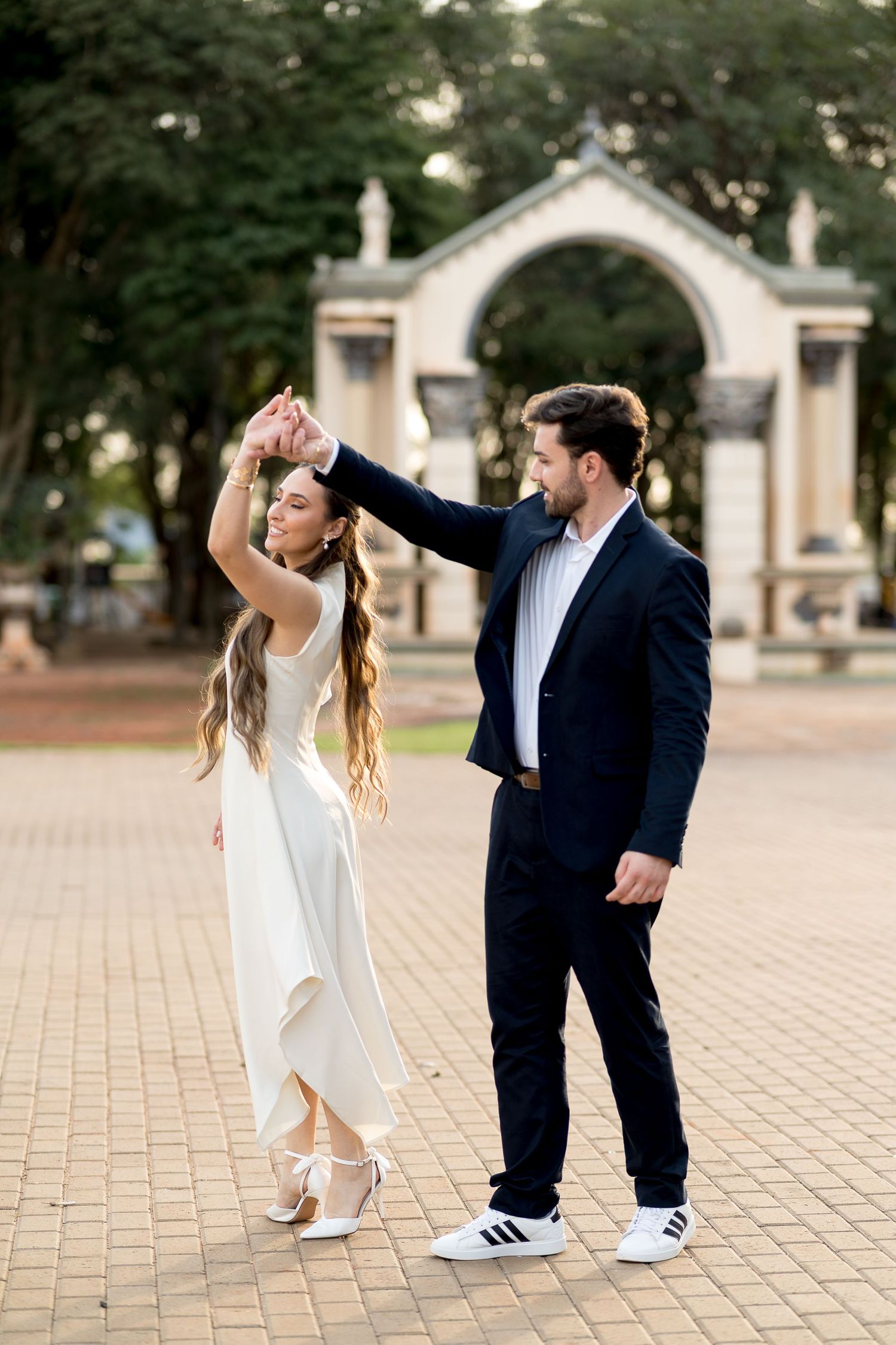 Couple dancing outdoors near an arch, woman in white dress spinning, man in dark suit, park setting.